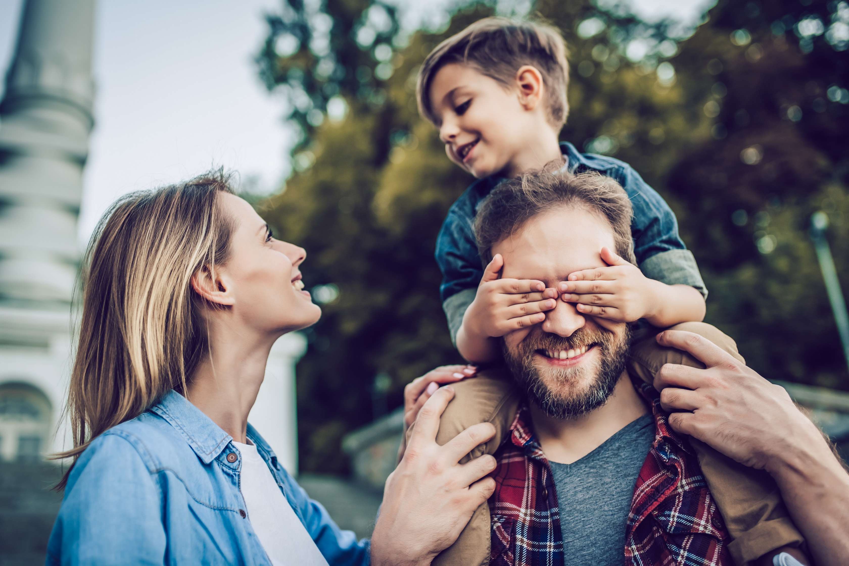 Allianz Gesundheitswelt - ElternCoach mit STEP: ein Kind im Jeanshemd sitzt auf den Schultern eines Manns und hält diesem die Augen zu. Der Mann trägt ein karriertes Hemd. Links daneben steht eine Frau und lacht das Kind an.