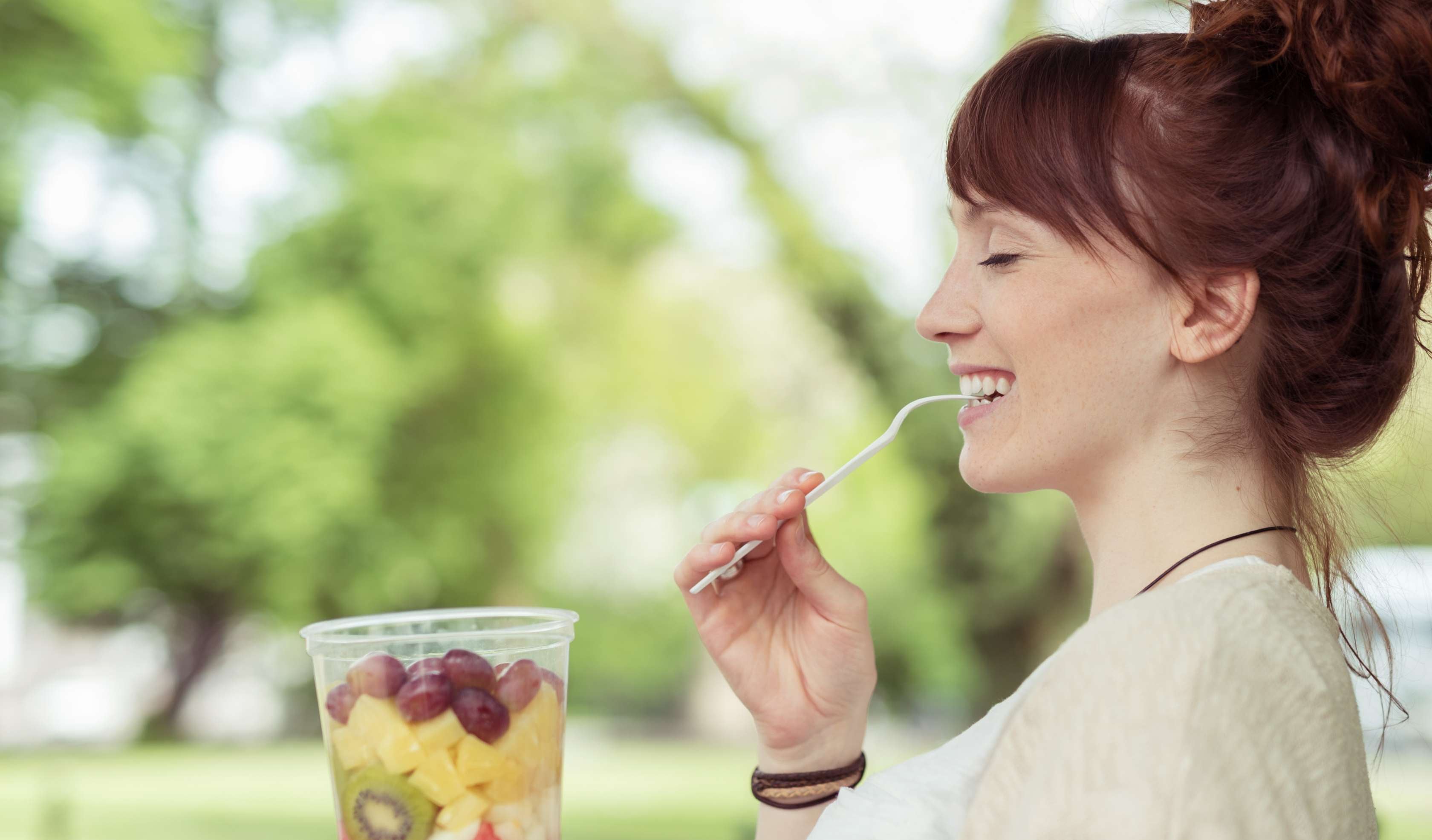 Allianz Gesundheitswelt - Achtsames Essen: eine Frau mit Dutt sitzt auf einer Parkbank und isst einen Becher mit Obst.