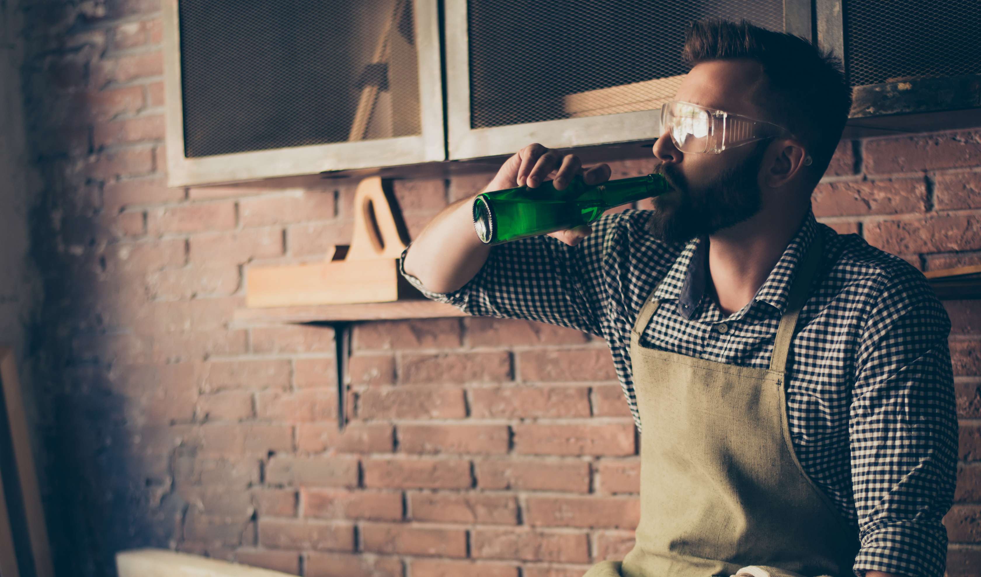 Allianz Geundheitswelt - Alkohol am Arbeitsplatz: Ein Mann mit Lederschürze lehnt sich an eine Holzwerkbank und  trinkt Bier aus einer grünen Glasflasche.
