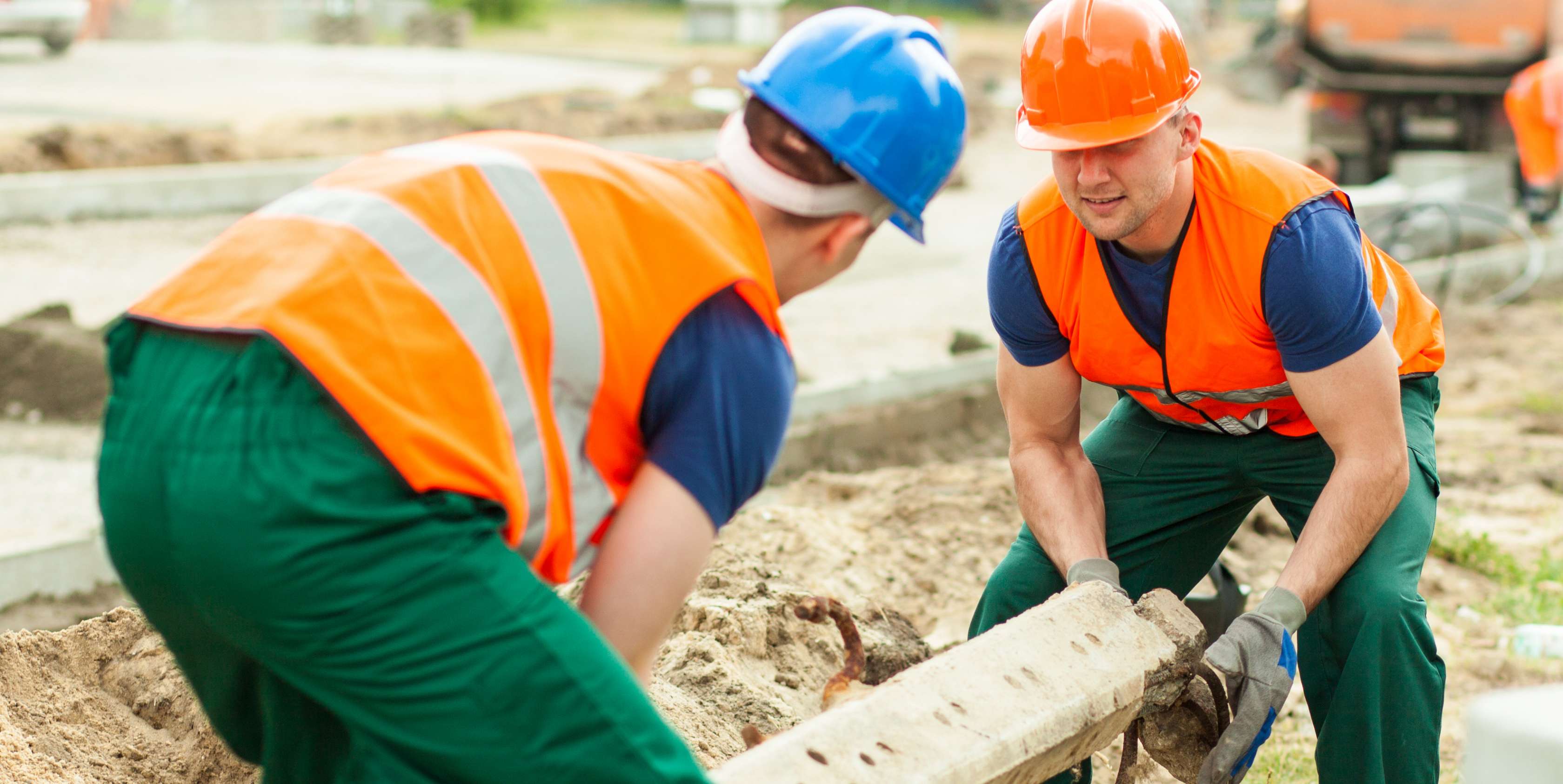 Allianz Gesundheitswelt - Bücken, Heben, Tragen: zwei Bauarbeiter mit orangenen Warnwesten und Schutzhelmen tragen einen Betonpfeiler.