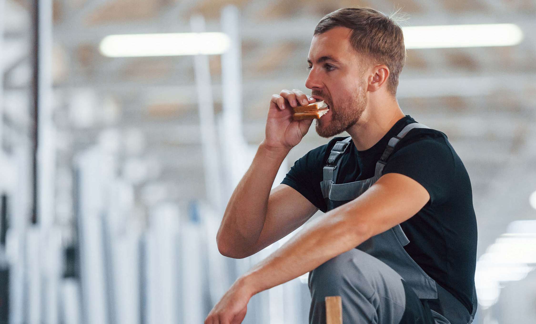 Allianz Gesundheitswelt - Ernährung Nachtschicht: ein junger Mann mit Latzhose isst während seiner Pause in der Nachtschickt ein Sandwich.