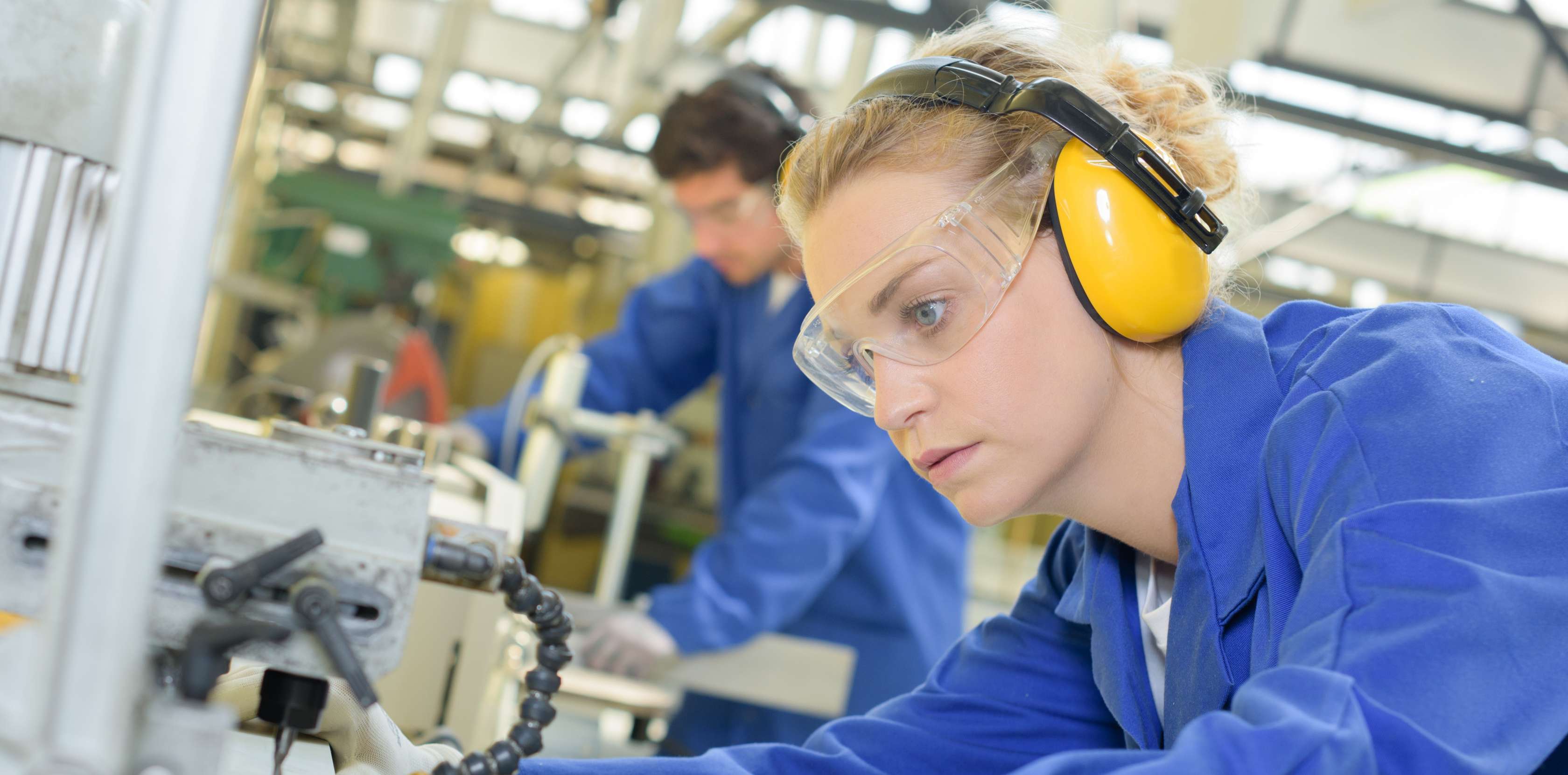 Allianz Gesundheitswelt - Langes Stehen am Arbeitsplatz: Eine junge Frau mit Schutzkleidung - Blauem Schutzanzug, Handschuhen und Ohrenschutz - steht an einer Maschine für Metalle.