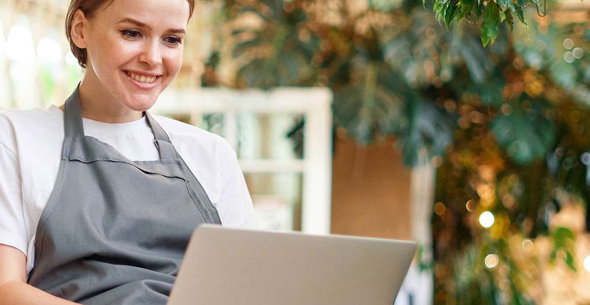 Allianz Gesundheitswelt - Luftfeuchte am Arbeitsplatz: eine junge Frau mit Schürze arbeitet an ihrem Laptop. Im Hintergrund sind Pflanzen zu erkennen.