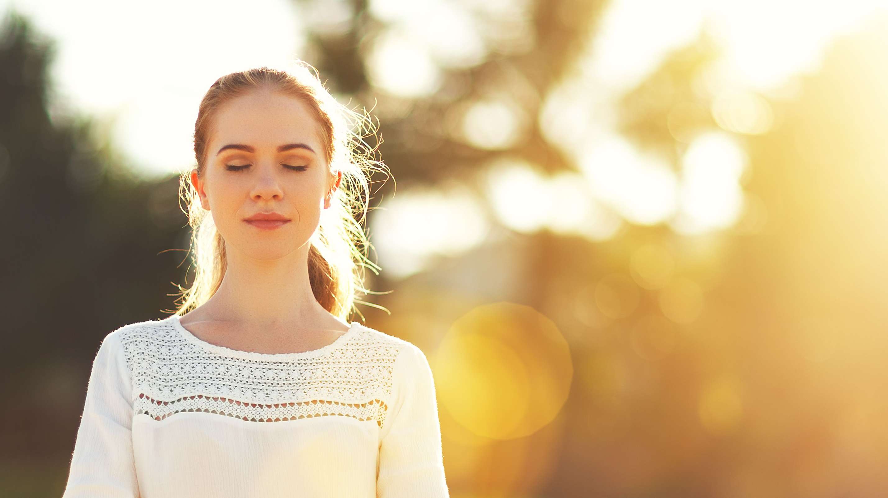 Allianz Gesundheitswelt - Entspannungsmethoden Test: eine junge Frau mit mittelhohem Zopf sitzt im Schneider sitz am Strand und meditiert. Im Hintergrund geht zwischen den Bäumen die Sonne auf.
