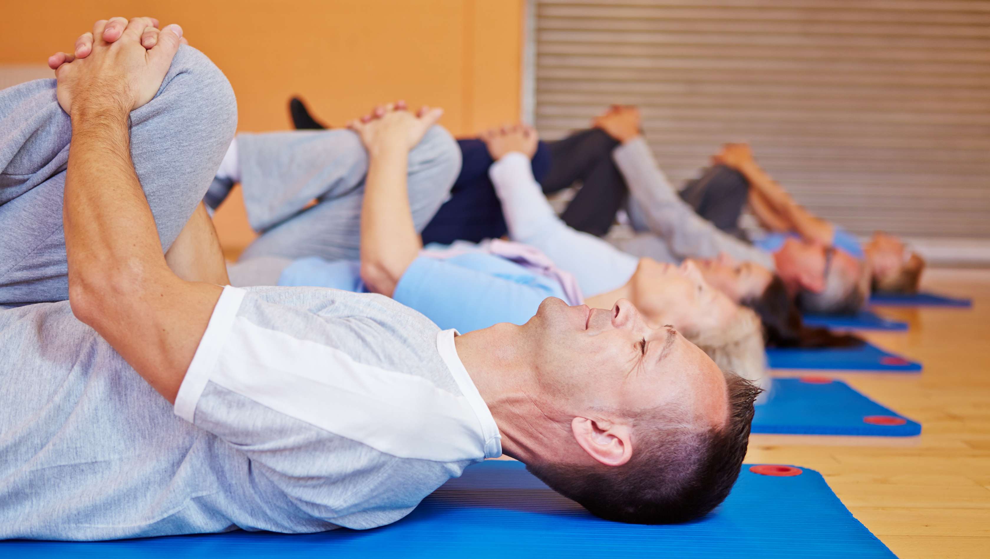 Allianz Gesundheitswelt - Rückenprogramm Mobilisation: eine Gruppe von Menschen liegt in einer Turnhalle auf Fitnessmatten und zieht ein Knie in Richtung Kinn, um den Rücken zu entlasten.