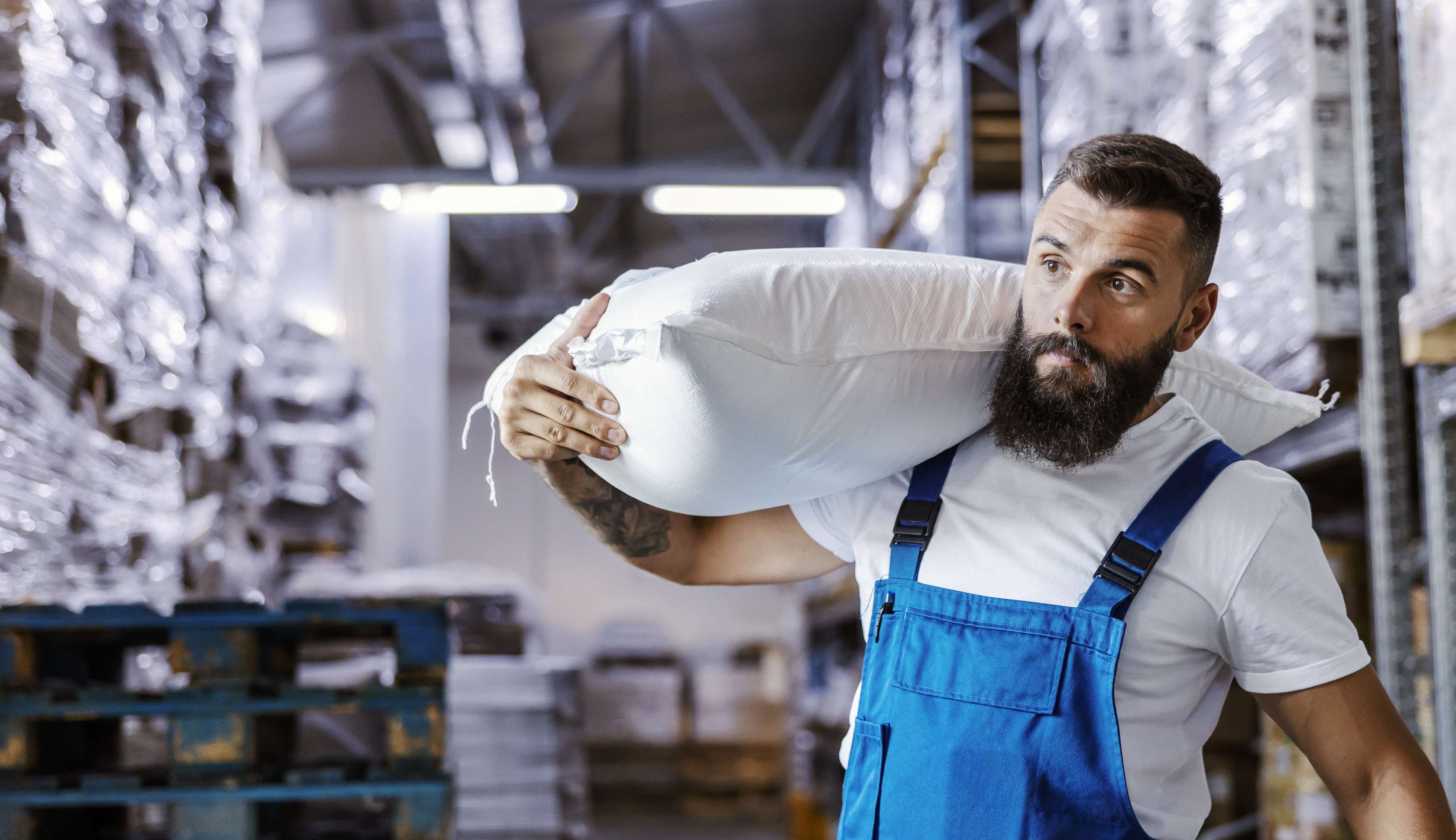 Allianz Gesundheitswelt - Schwere Lasten am Arbeitsplatz: Ein Mann mit blauer Latz-Arbeitshose trägt einen schweren Betonsack durch das Lager.