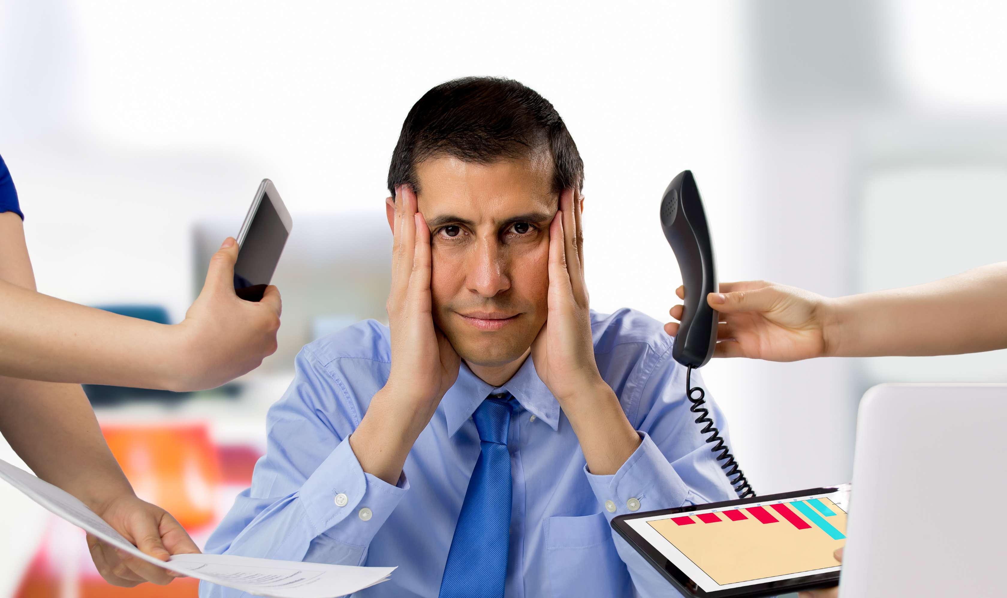 Allianz Gesundheitswelt - Stressauslöser: ein Mann mit blauem Hemd und blauer Krawatte sitzt frustriert an einem weißen Schreibtisch. Von links und rechts werden Telefonhörer, Tablet und Smartphone herangereicht.