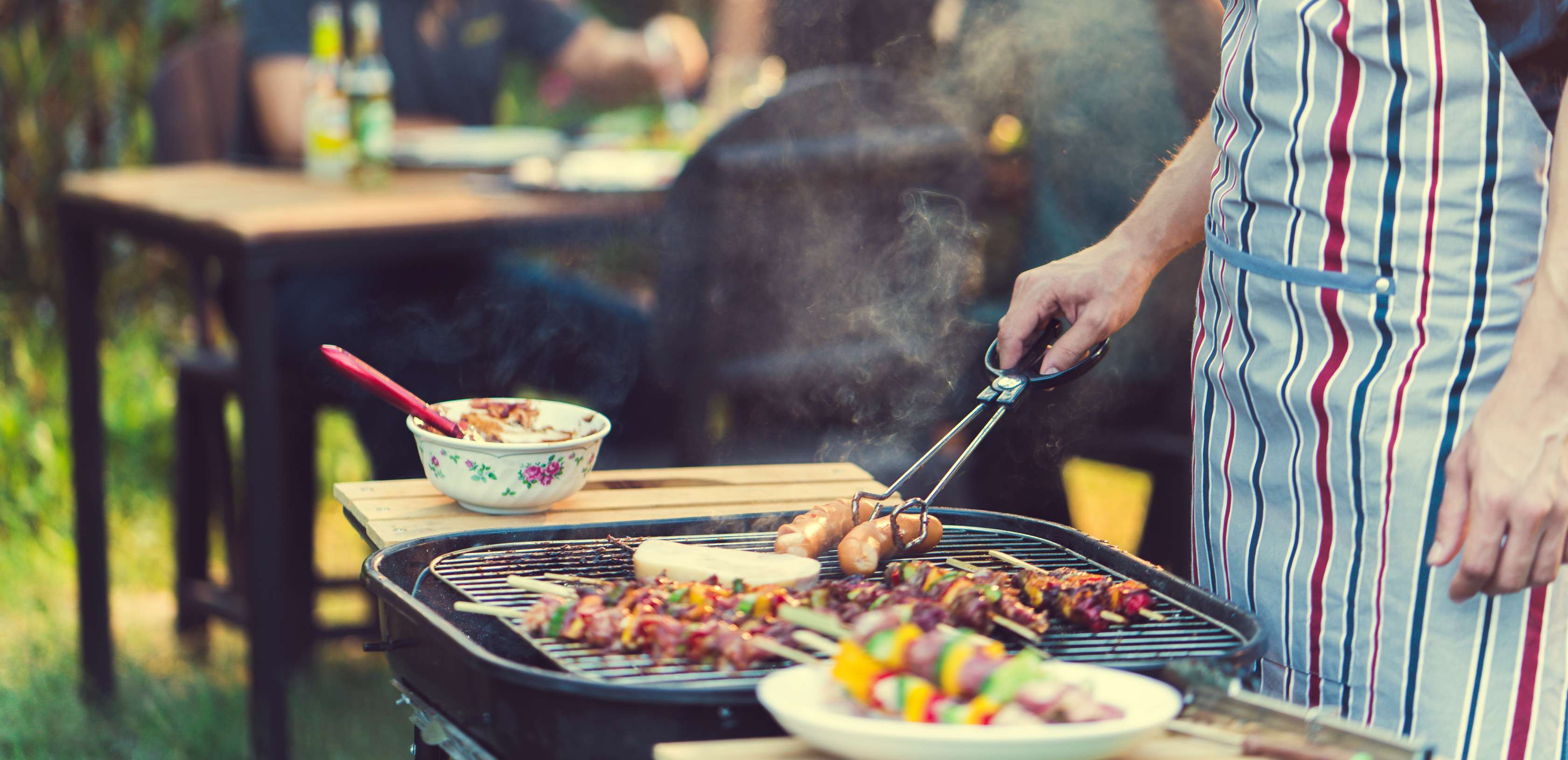 Allianz Gesundheitswelt - Grilltipps: eine Person steht am Grill und grillt Fleisch und Würste. Daneben steht eine Gemüseplatte.