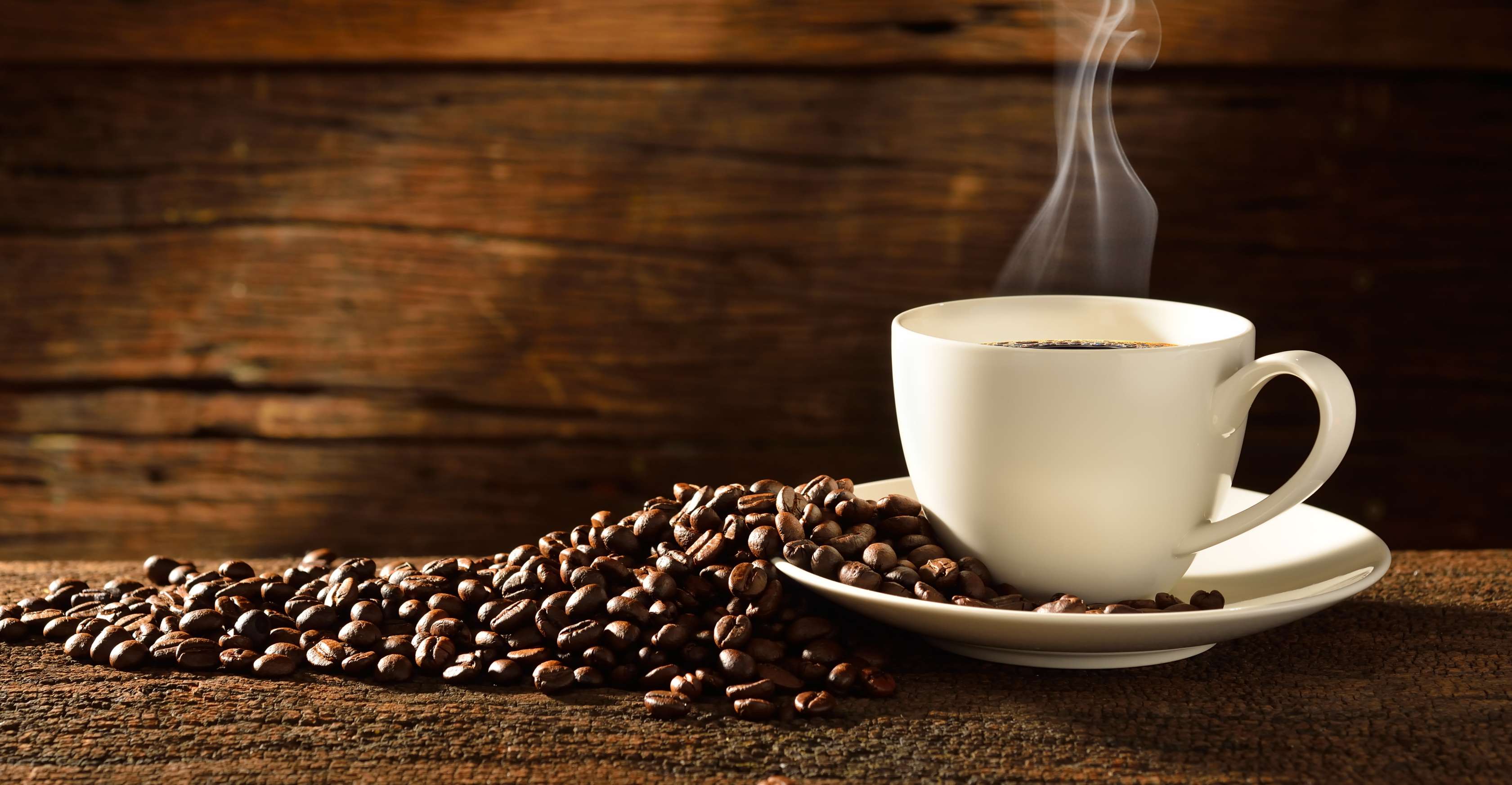 Allianz Gesundheitswelt - Fakten Kaffee: eine weiße Kaffeetasse steht auf einem dunklen Holzuntergrund. Um die Tasse sind Kaffeebohnen verteilt.