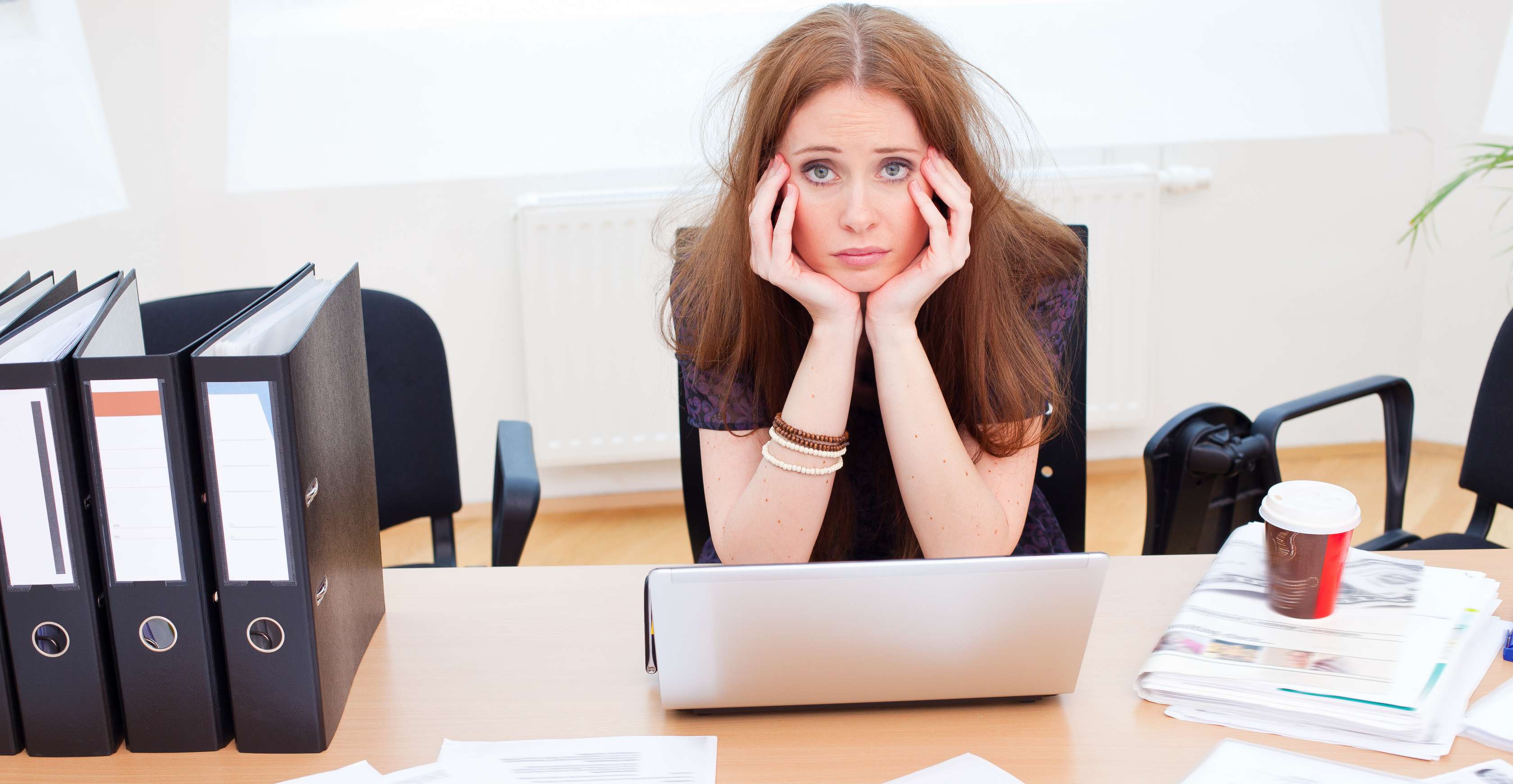 Allianz Gesundheitswelt - Zeitmanagement im Job: Eine junge Frau mit langen Haaren sitzt frustriert am Schreibtisch vor ihrem Laptop. Vor ihr sind Papierstapel ausgebreitet.