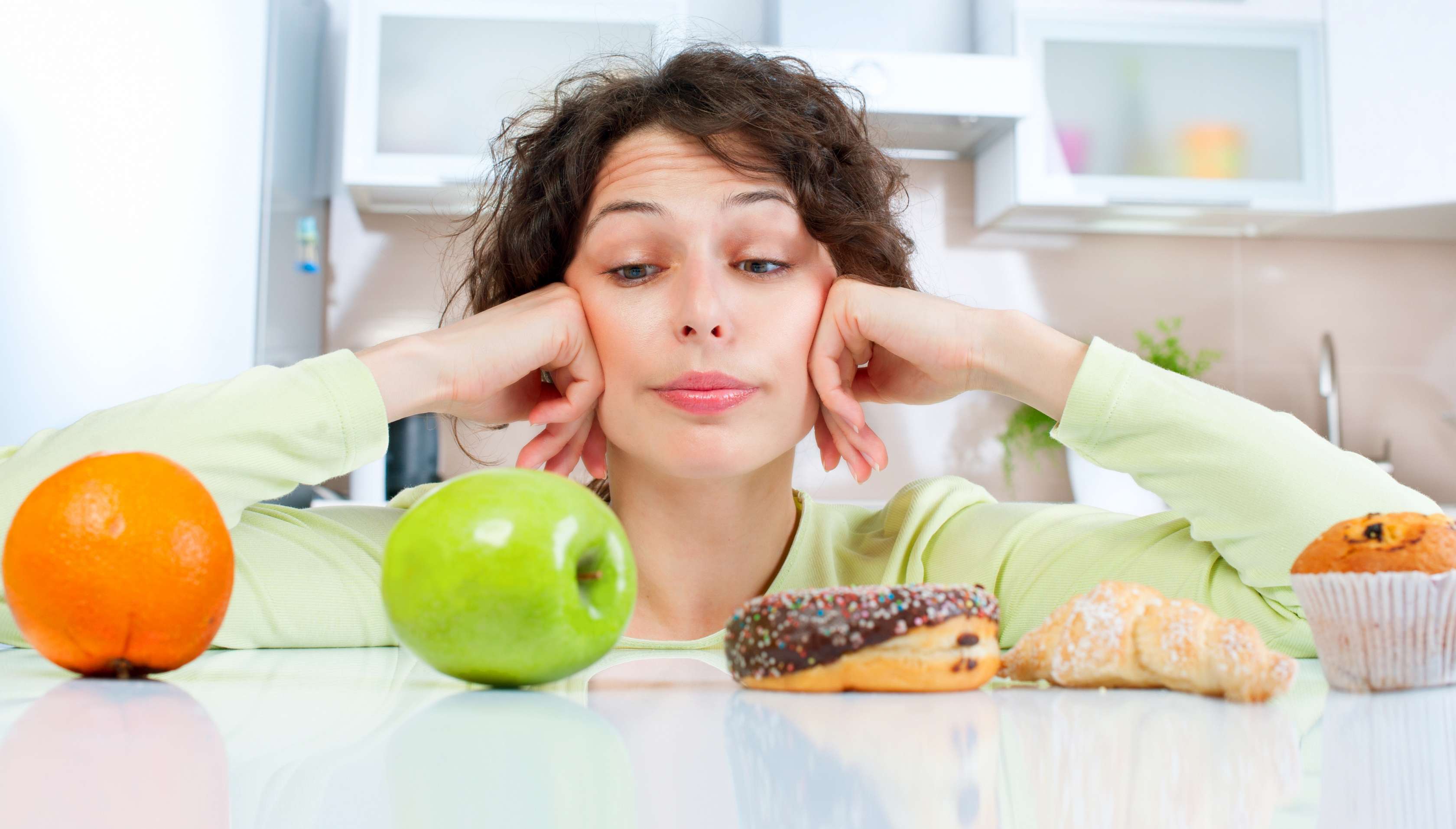 Allianz Gesundheitswelt - Abnehmtipps: eine Frau sitzt vor obst und süßen Teilchen und betrachtet den grünen Apfel skeptisch.