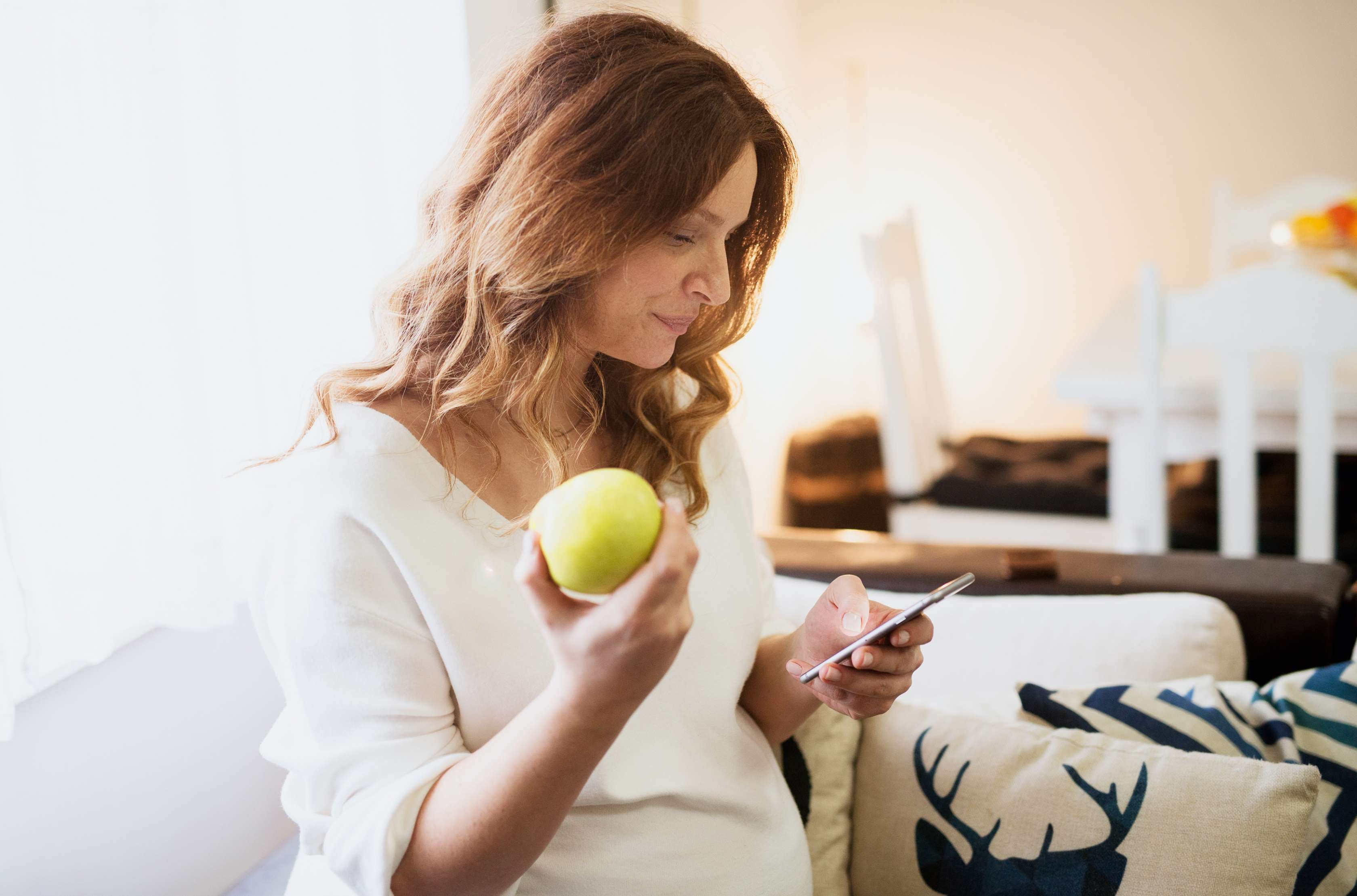 Allianz Gesundheitswelt - Komplikationen in der Schwangerschaft: eine Frau sitzt auf der Couch und sieht auf ihr Smartphone. Sie informiert sich über Risiken in der Schwangerschaft. in der anderen Hand hält sie einen Apfel, von dem sie bereits abgebissen hat.