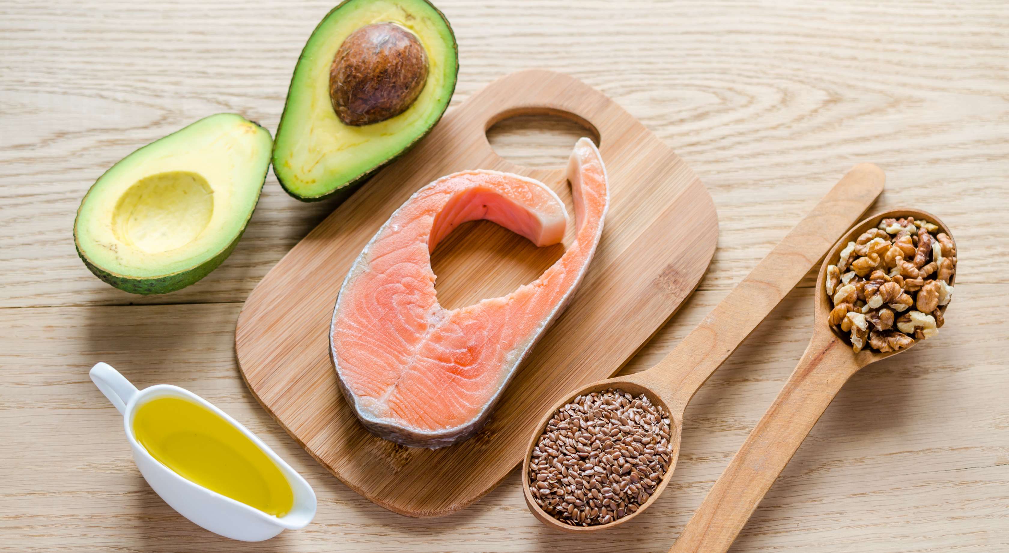 Allianz Gesundheitswelt - gutes Fett, böses Fett: auf einer hellen Holzarbeitsplatte liegen Avocados, ein Kännchen mit Öl, ein Brett mit Tunfisch und zwei Löffel mit Nüssen und Leinsamen..