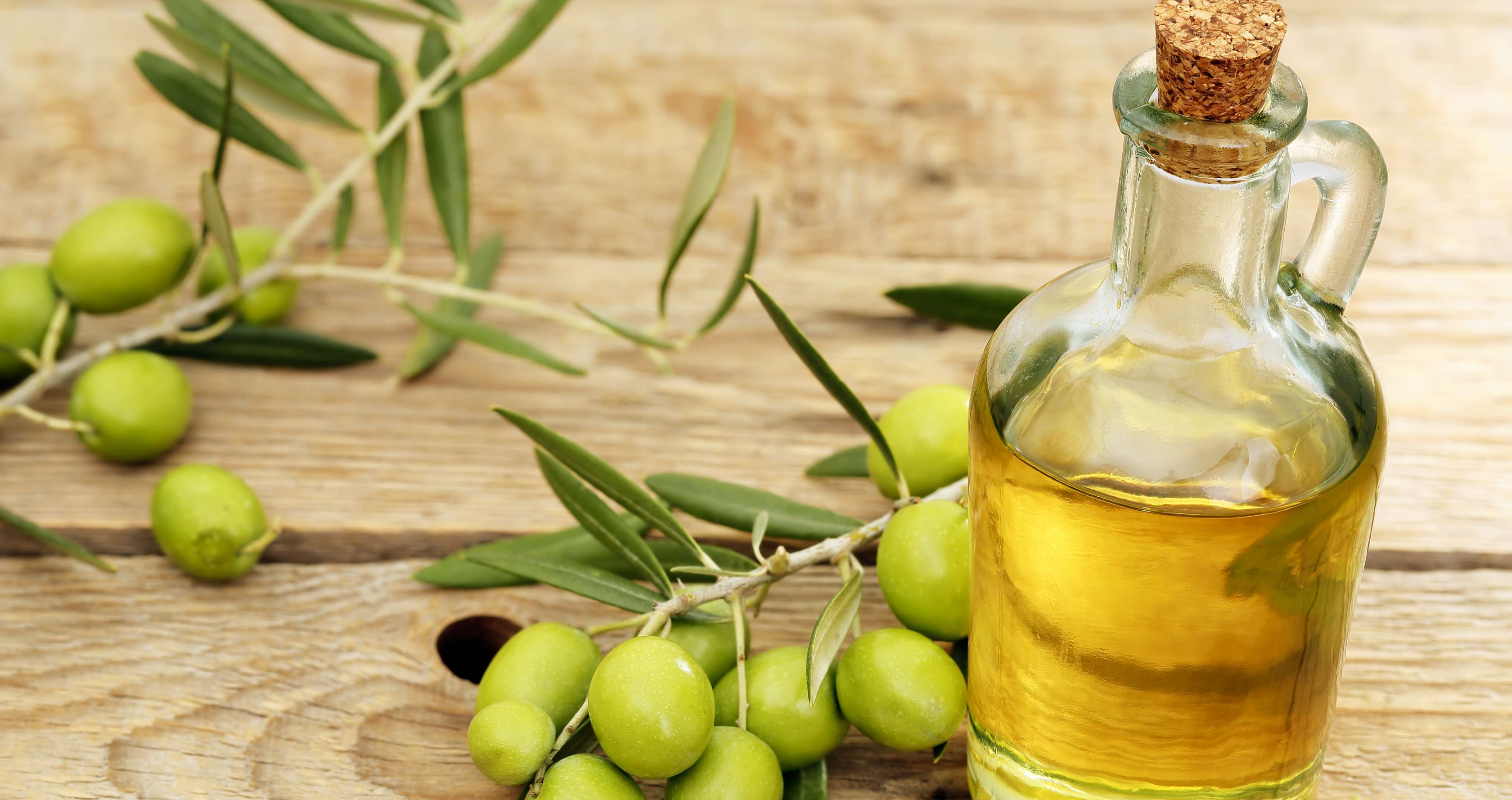 Allianz Gesundheitswelt - Olivenöl: eine Karaffe mit Olivenöl steht  auf einem Holzuntergrund. Daneben liegen Olivenzweige.