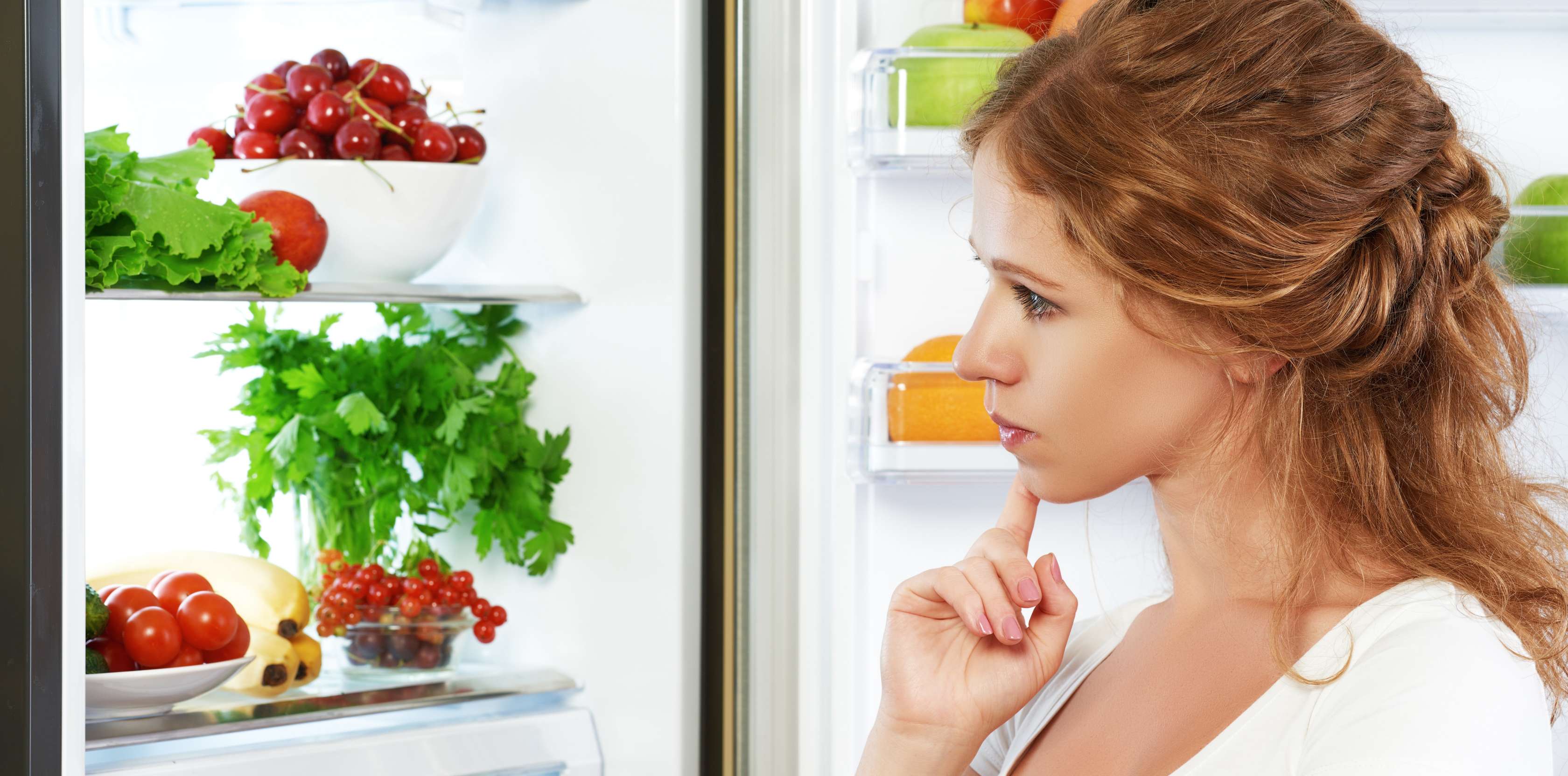 Allianz Gesundheitswelt - Orthorexie: eine junge frau mit gelockten Haaren steht vor dem geöffneten Kühlschrank und betrachtet skeptisch den Inhalt.