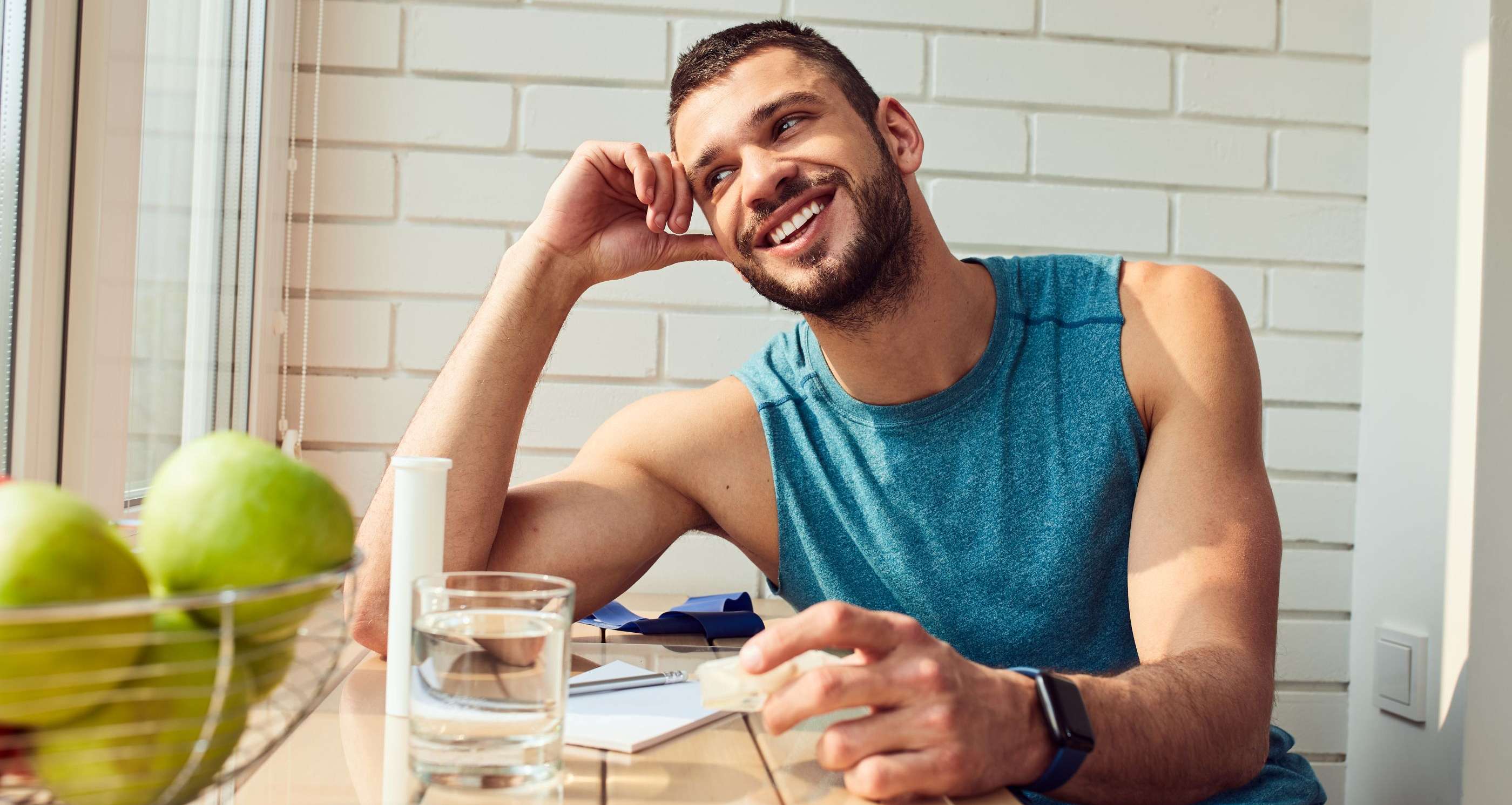 Allianz Gesundheitswelt - Vitamin C: Ein lächelnder Mann sitzt an einem Tisch mit Obstschale und Wasserglas und schaut aus dem Fenster. 