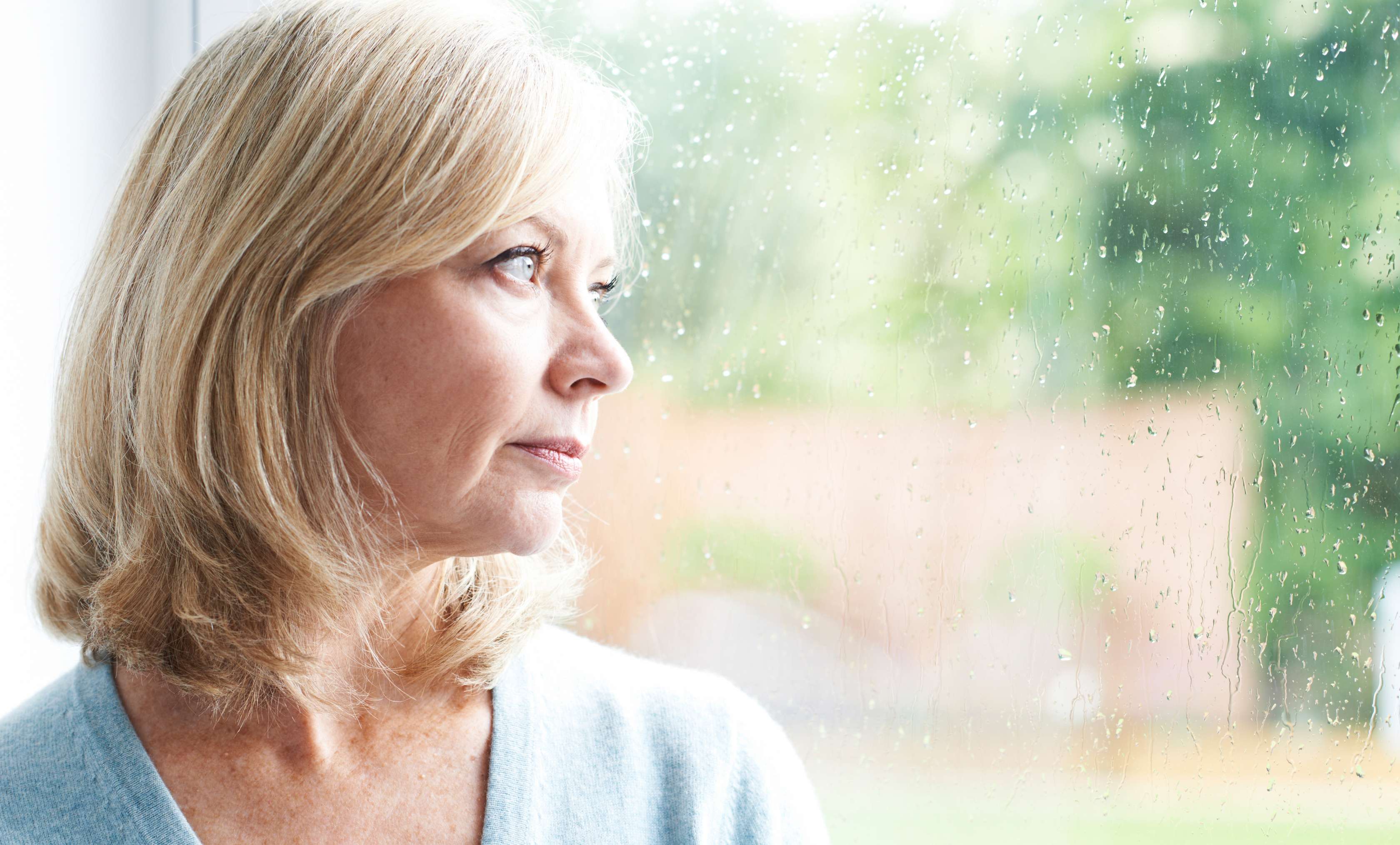 Allianz Gesundheitswelt - Rückschläge überwinden: eine ältere Dame steht gedankenverloren am Fenster und sieht in die Ferne.