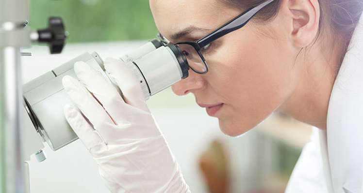 Allianz Gesundheitswelt - Behandlung: Eine Frau mit zusammengebundenen Haaren sieht in einem Labor durche in Mikroskop. Sie trägt einen Laborkittel und eine Brille.
