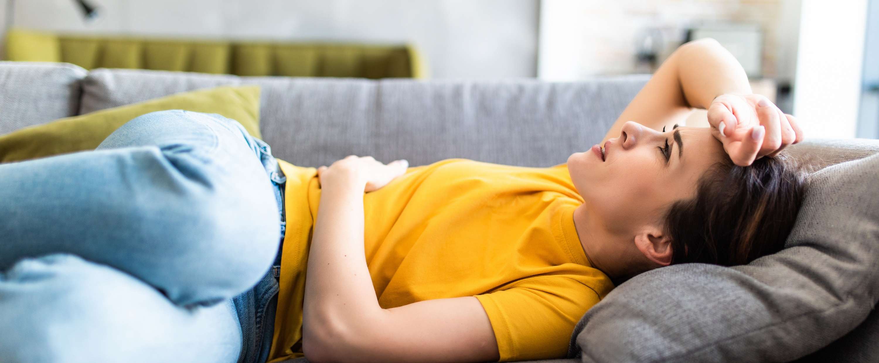 Allianz Gesundheitswelt - Was hilft gegen Kopfschmerzen: eine Frau liegt schmerzverzerrt auf dem Sofa und hält sich Bauch und Kopf