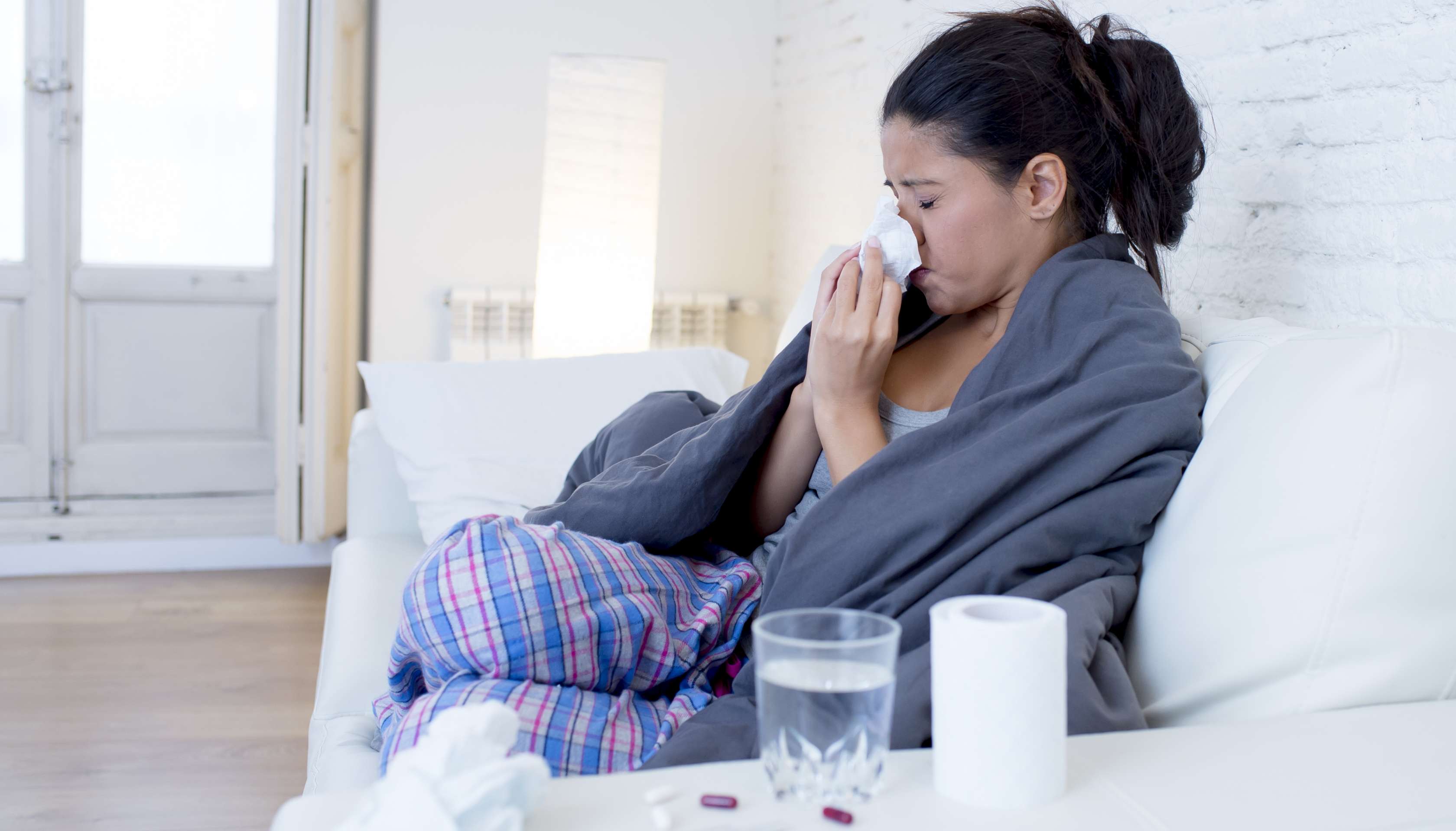 Allianz Gesundheitswelt - Schnupfen: eine junge Frau sitzt verschnupft und in eine Decke eingewickelt auf der Couch. Sie niest in ein Taschentuch.