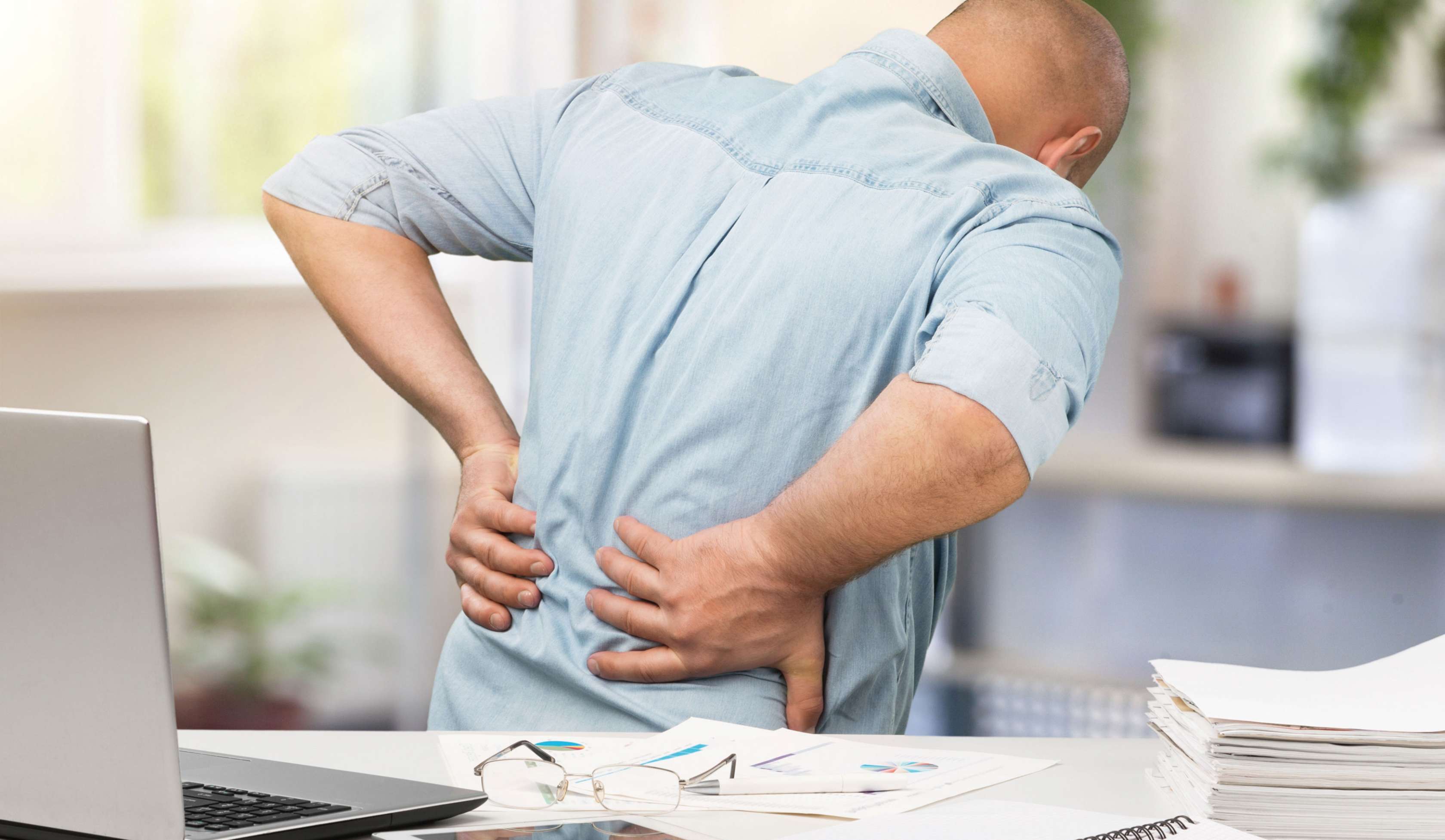 Allianz Gesundheitswelt - Rückenbeschwerden: ein Mann im Hemd steht mit dem Rücken vor einer Schreibtischpaltte. Seine Hände stemmt er in den unteren Rücken, um seine Rückenschmerzen zu lindern.