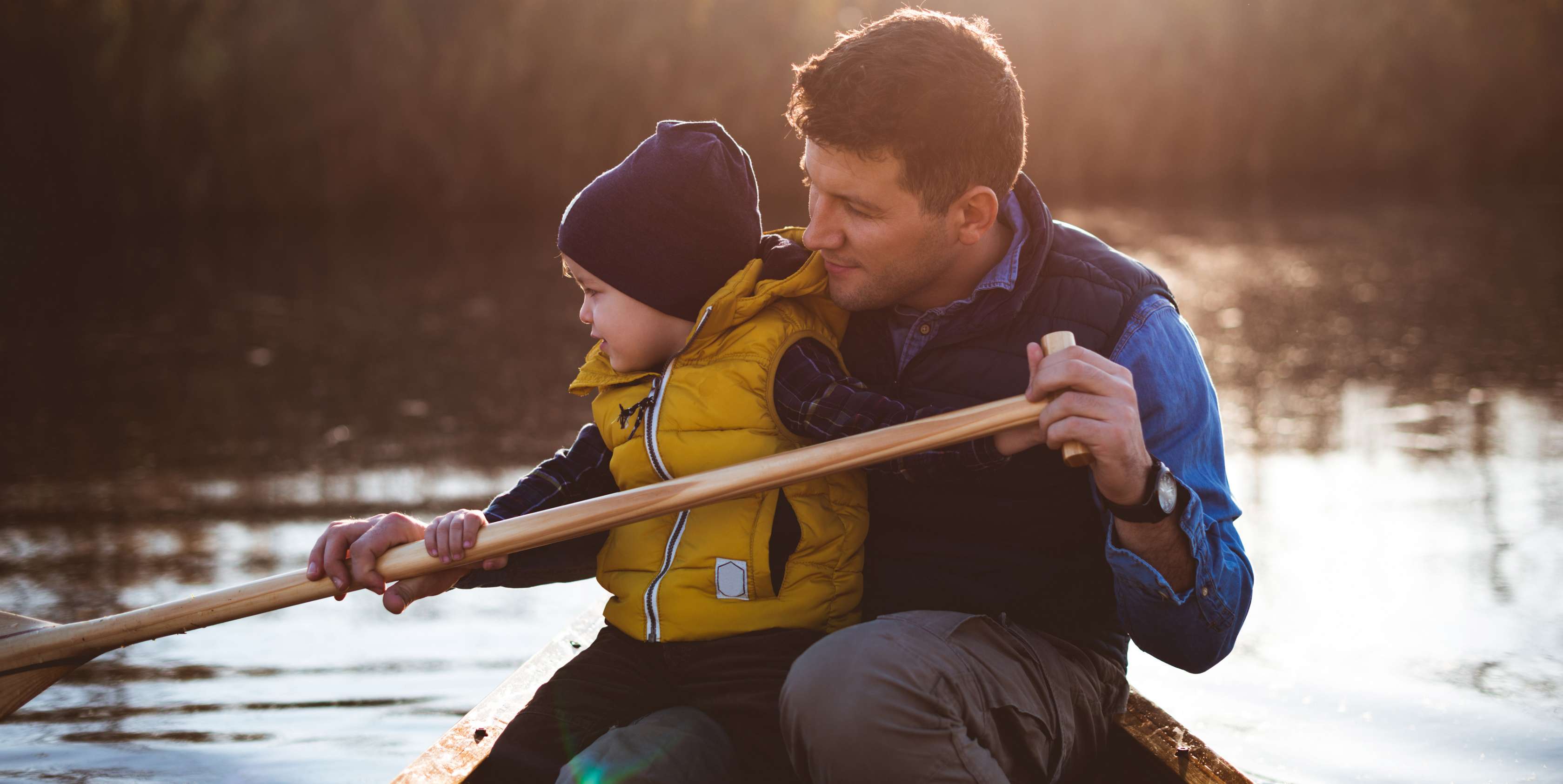 Allianz Gesundheitswelt - was ist Rheuma: ein Vater mit schwarzer Steppweste und sein Kind sitzen in einem Ruderboot auf einem See. Durch die feuchte Luft können rheumatische Schmerzen zutage gefördert werden.