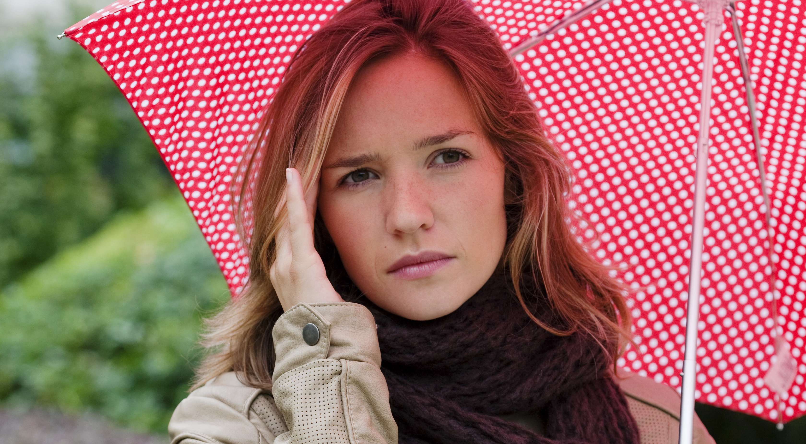 Allianz Gsesundheitswelt - Wetterfühligkeit: eine junge Frau steht unter ihrem Regenschirm umd massiert sich mit einer Hand ihre Stirn, um ihre Wetterfühligkeit zu mindern.