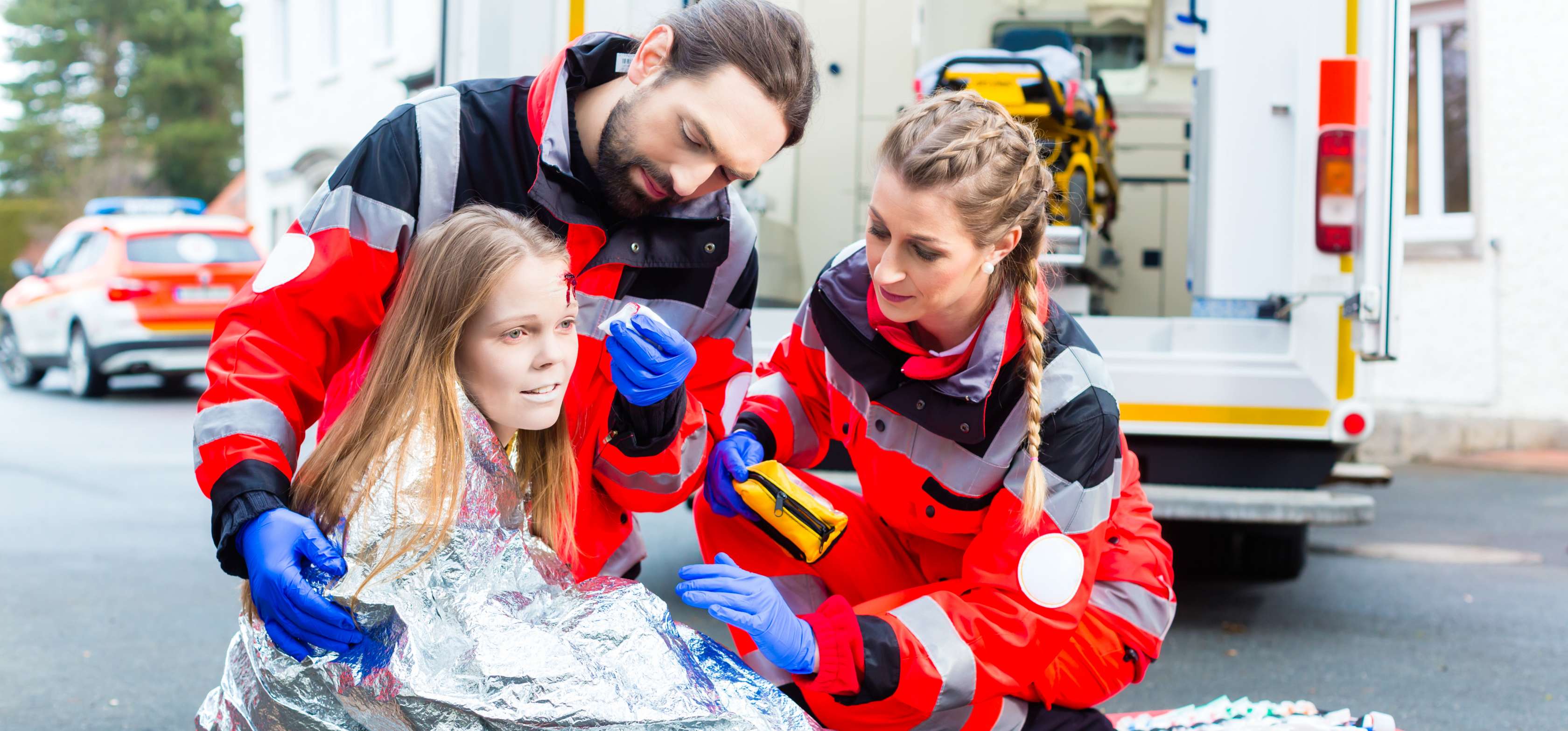 Allianz Gesundheitswelt - Erste Hilfe: eine junge Frau wird nach einem Unfall von einem Sanitäter und einer Sanitäterin behandelt. Die Frau sitzt mit einer Platzwunde und eingewickelt in einer Folie auf einer Trage.