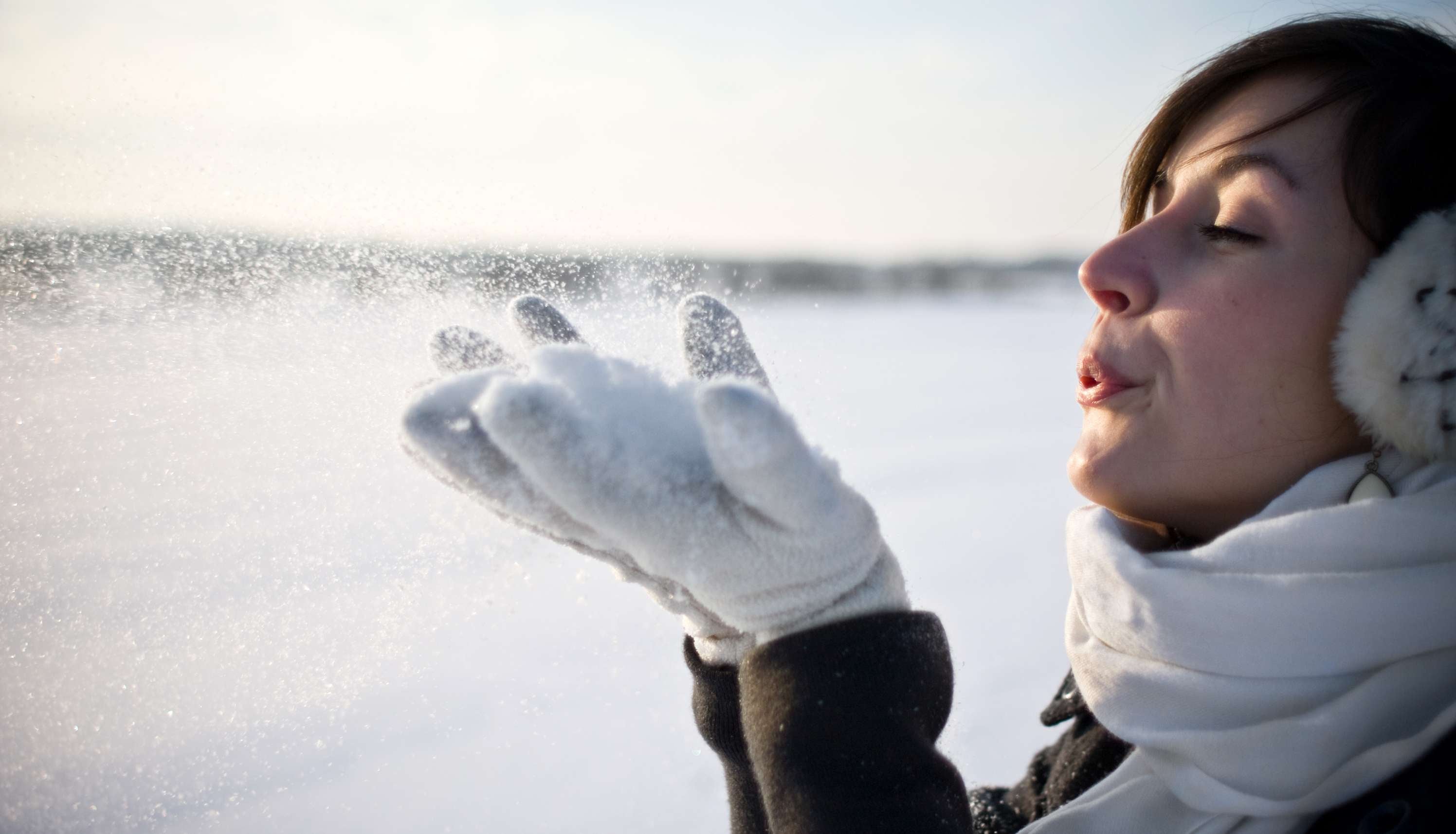 Allianz Gesundheitswelt - Hautpflege im Winter : eine Frau steht in einer Winterlandschaft und pustet Schnee aus ihren Händen in die Luft.