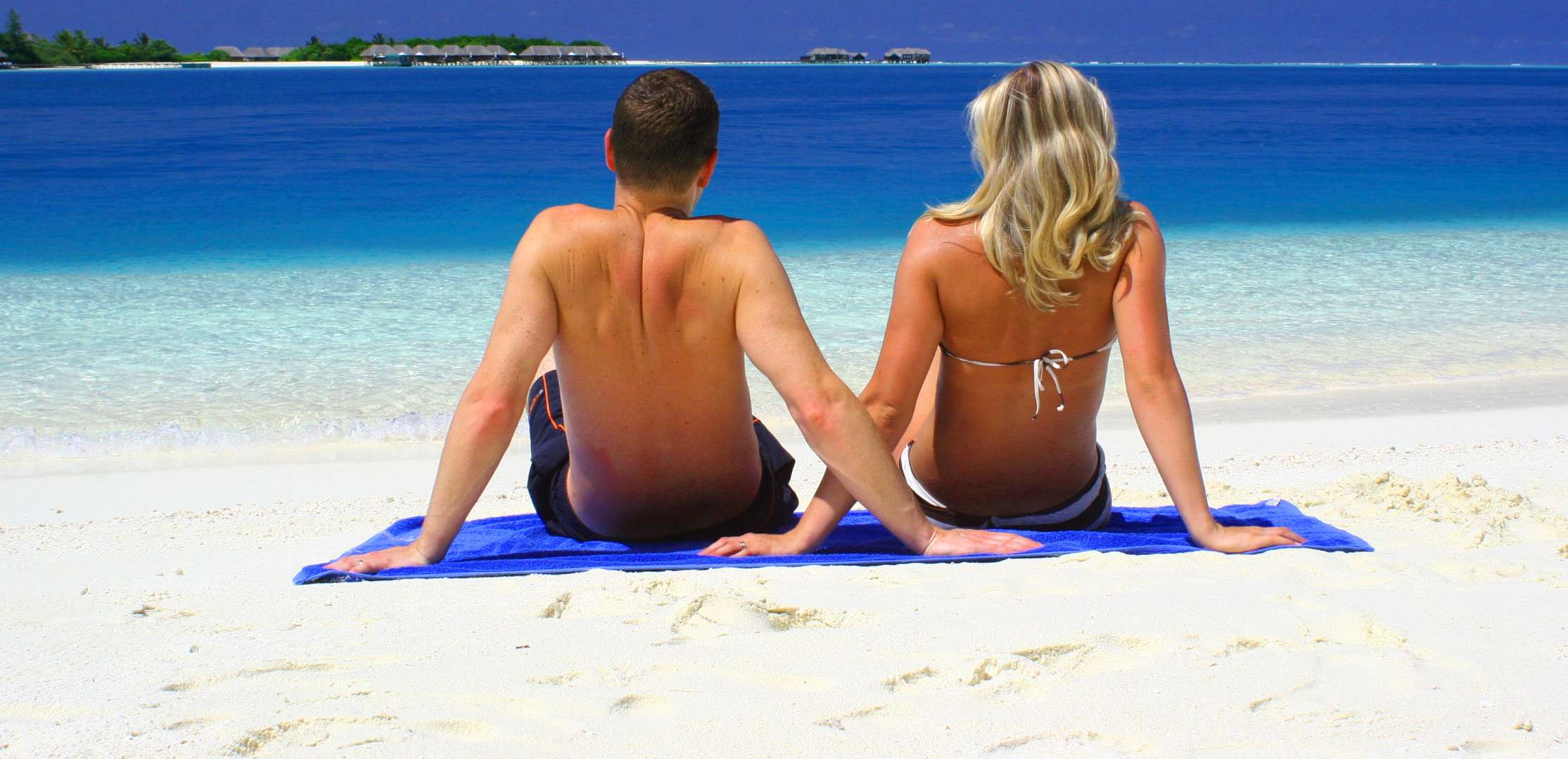 Allianz Gesundheitswelt - Hauttypen: ein Paar sitzt in Badehose und Bikini am Strand und blockt auf das mneer hinaus.