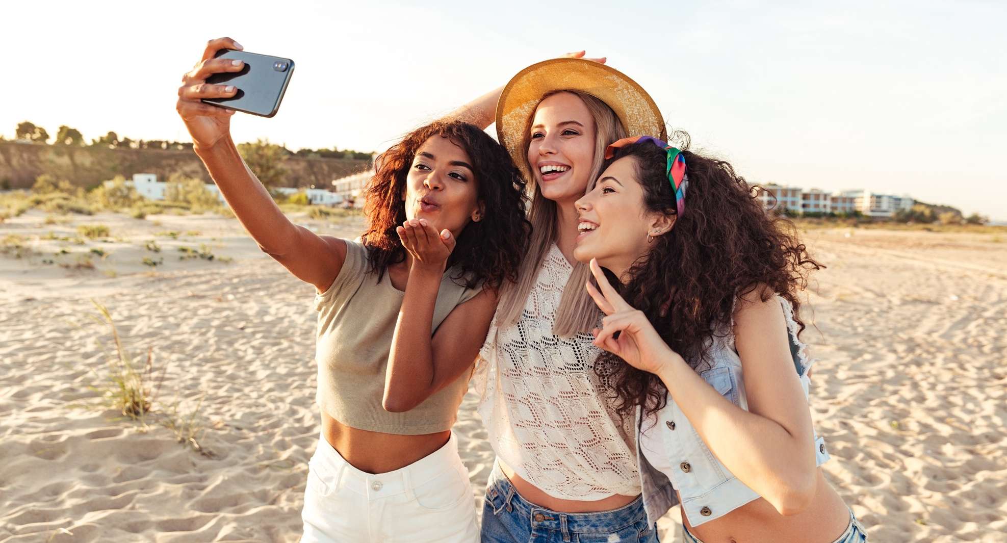 Allianz Gesundheitswelt - UV-Index: Drei junge Frauen lächeln am Strand in eine Handykamera