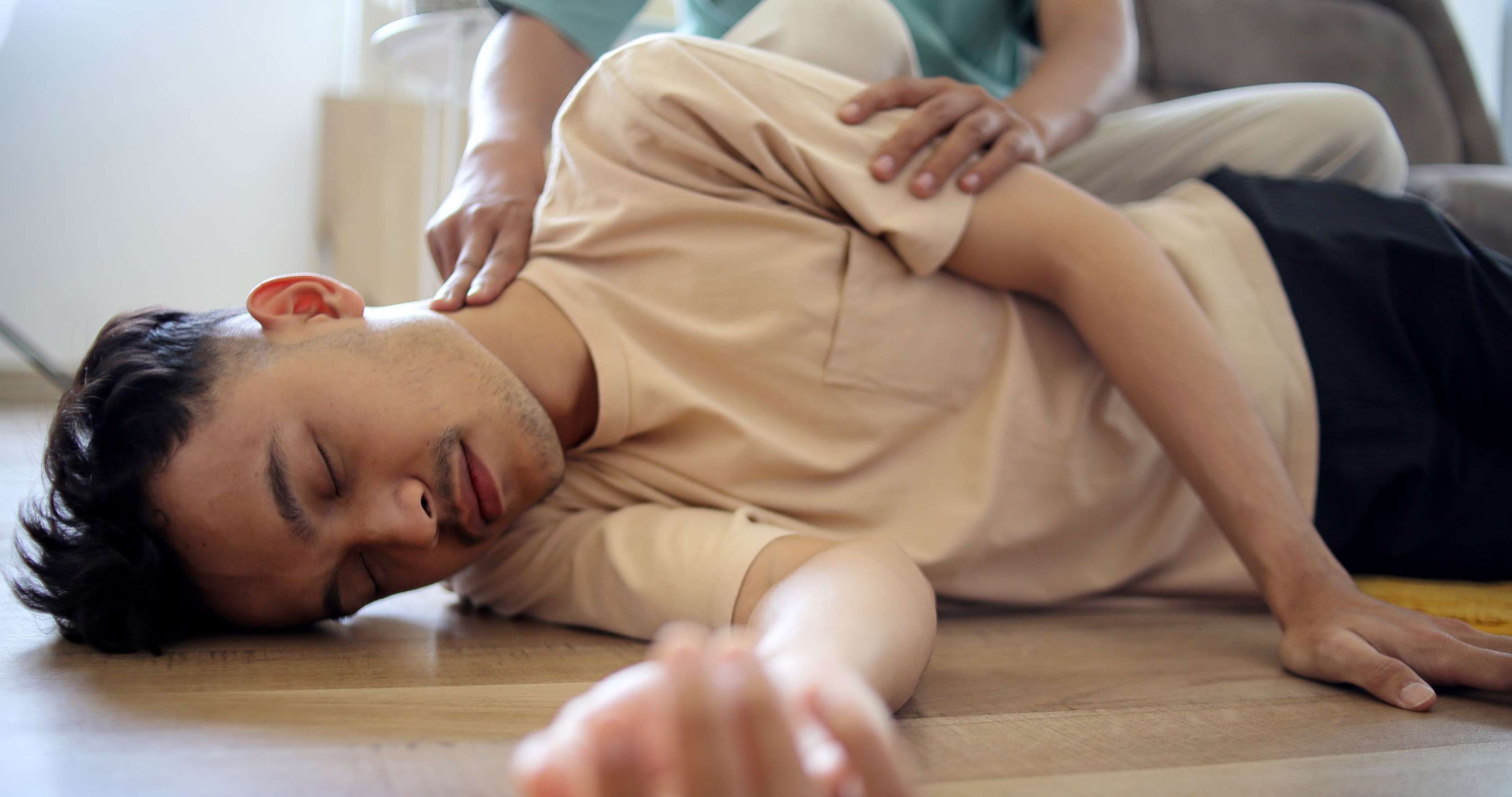 Allianz Gesundheitswelt - Erste Hilfe: eine bewusstlose Person liegt in der stabilen Seitenlage auf dem Boden, eine zweite Person fühlt ihren Puls am Hals.
