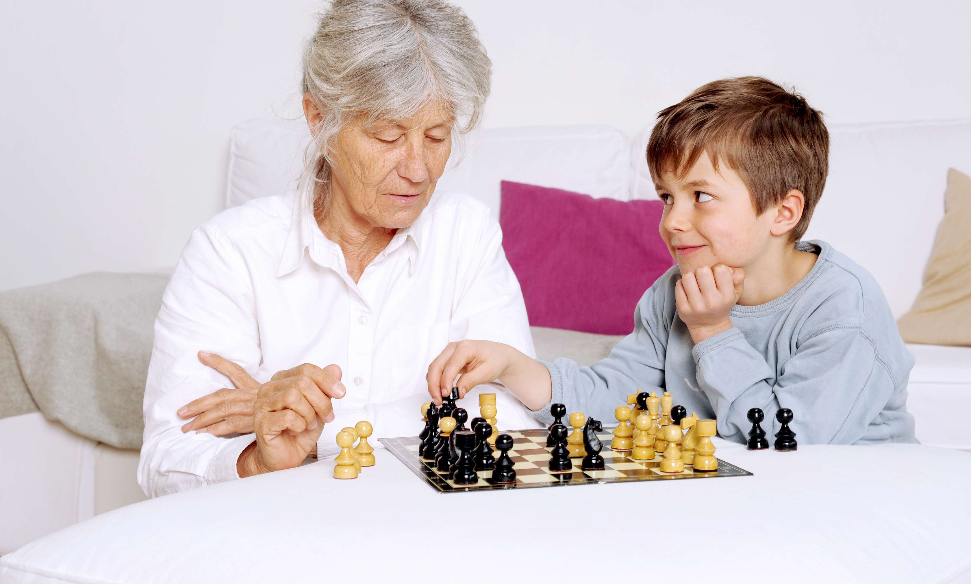 Allianz Gesundheitswelt - geistig fit bleiben: eine ältere Dame in weißer Bluse spielt mit ihrem Enkel in grauem Sweater Schach.