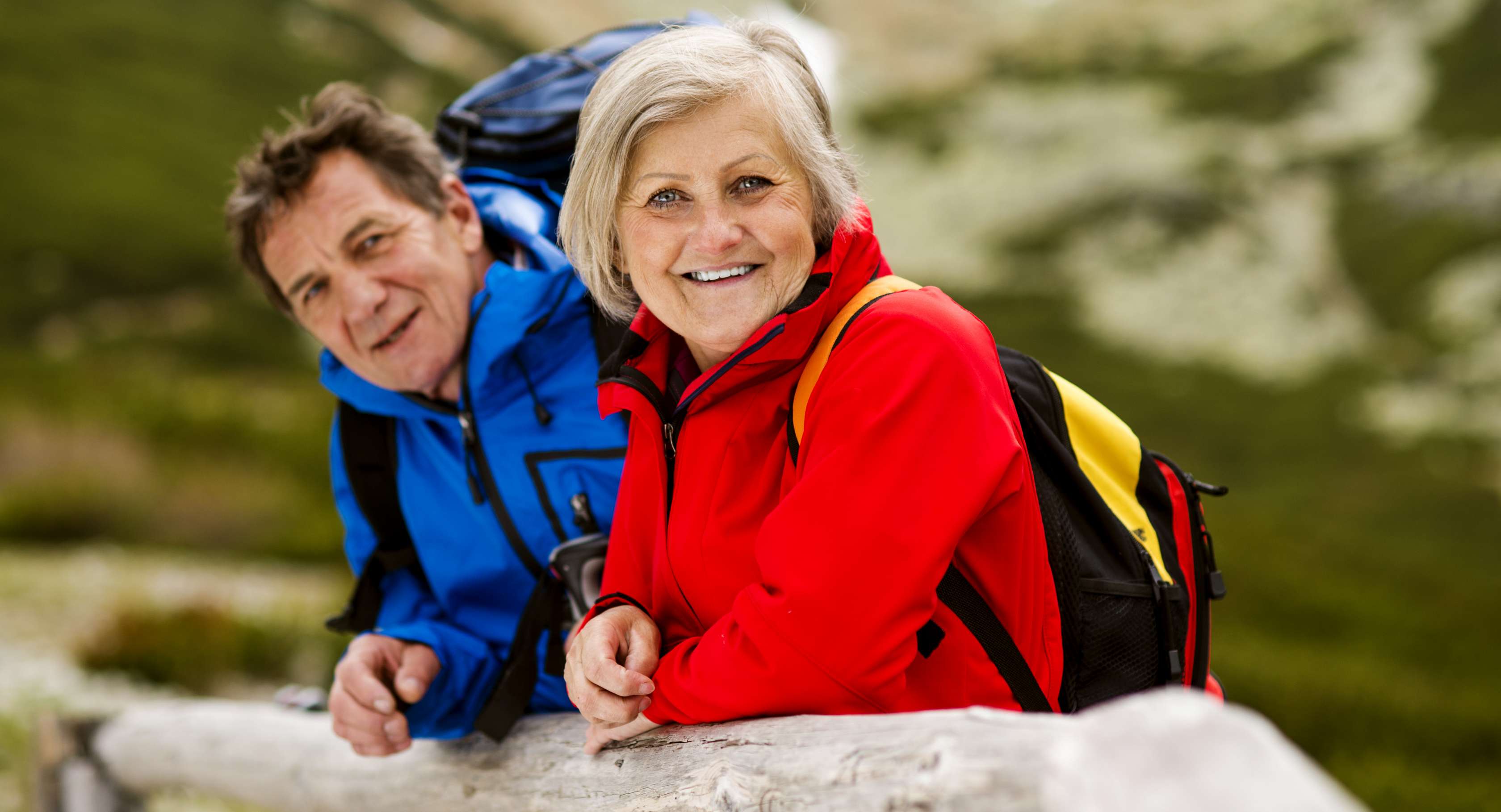 Allianz Gesundheitswelt - Mobilität erhalten: eub äöälteres Paar macht eine Pause vom Wandern. Beide tragenm Ourdoorjacken - die Frau eine rote und der Mann eine blaue - und einen Rucksack auf dem Rücken.