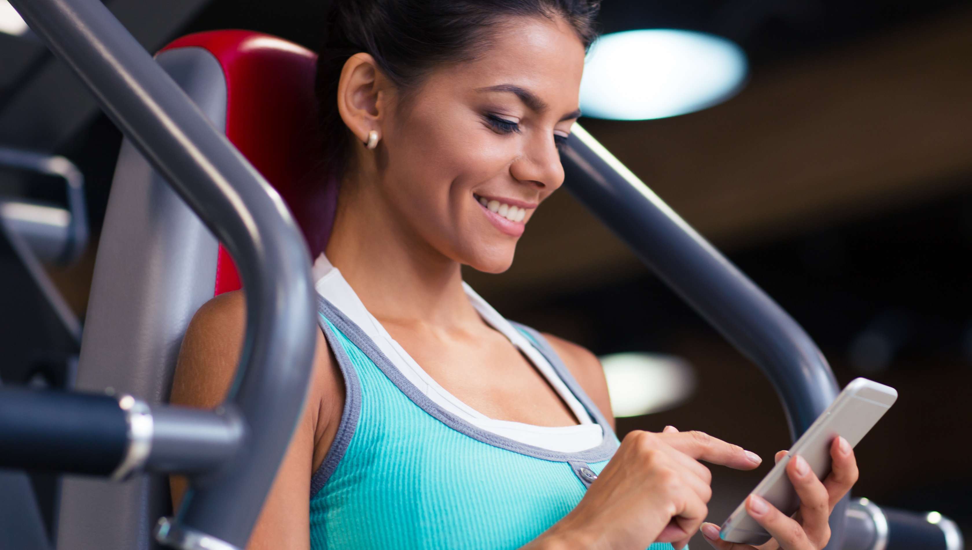 Allianz Gesundheitswelt - gesünder mit Apps: eine junge Frau sitzt auf einem Fitnessgerät im Fitnesstudio und tracktz ihren Trainingsfortschritt am Smartphone.