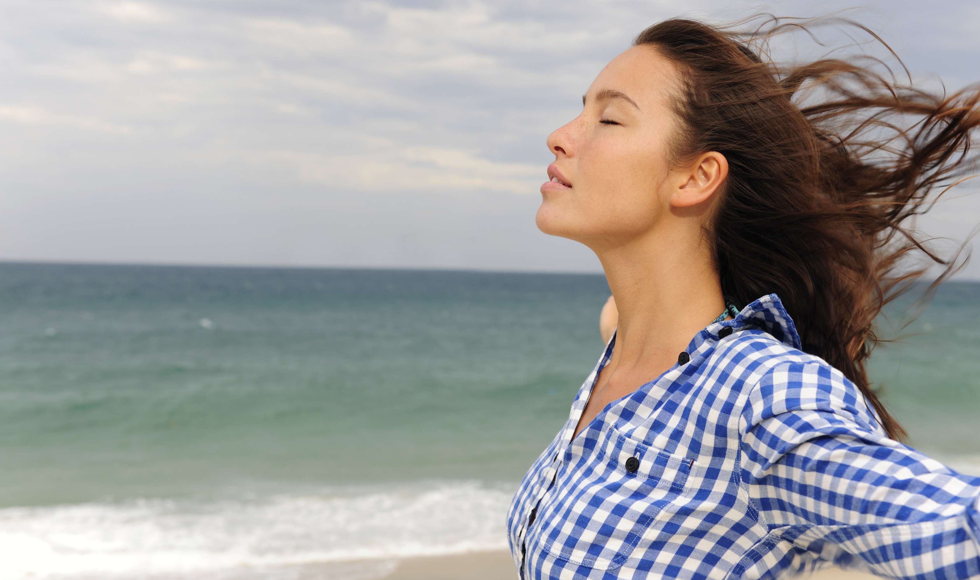Allianz Gesundheitswelt - Abstand gewinnen: eine Frau in einer blaukarierten Bluse steht mit ausgestreckten Armen am Meer und versucht den Kopf frei zu bekommen.