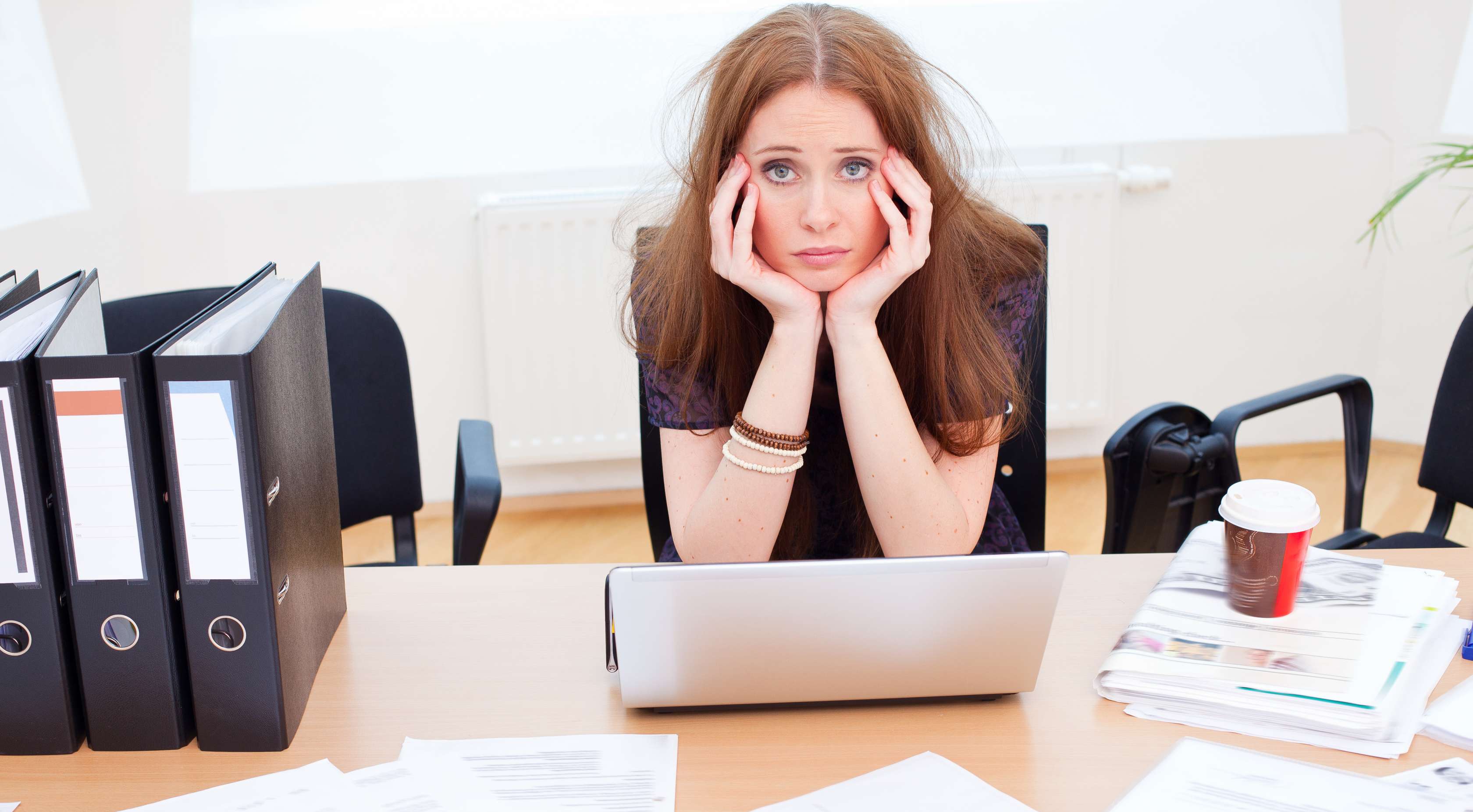 Allianz Gesundheitswelt - Burnout vorbeugen: eine junge Frau mit langen dunkelen Haaren und dunkelem kurzärmeligen Oberteil sitzt überfordert an ihrem chaotischem Schreibtisch.