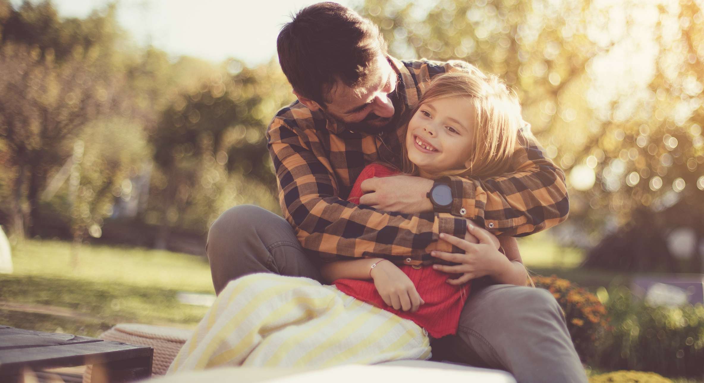 Allianz Gesundheitswelt - Nachhaltigkeit: Ein Mann im langärmligen Karohemd umarmt ein Kind, das in eine rote Decke eingewickelt ist, von hinten. Beide sitzen im Freien auf einer Sitzgruppe.