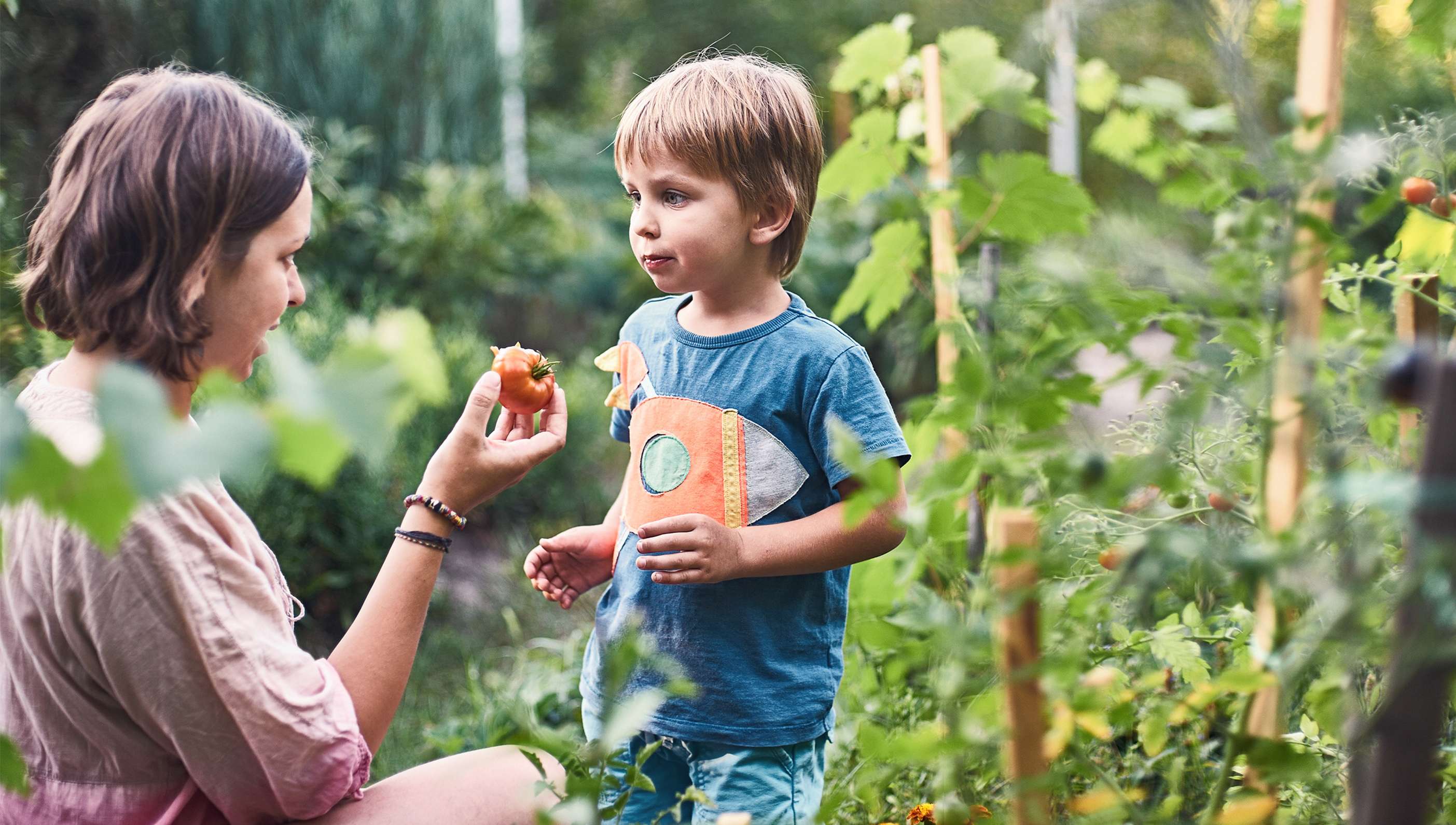 Allianz Gesungheitswelt - nachhaltige Kinderernährung: Eine Mutter lässt ihrem Kind zwischen verschiedenen Gemüsestauden eine Tomate kosten