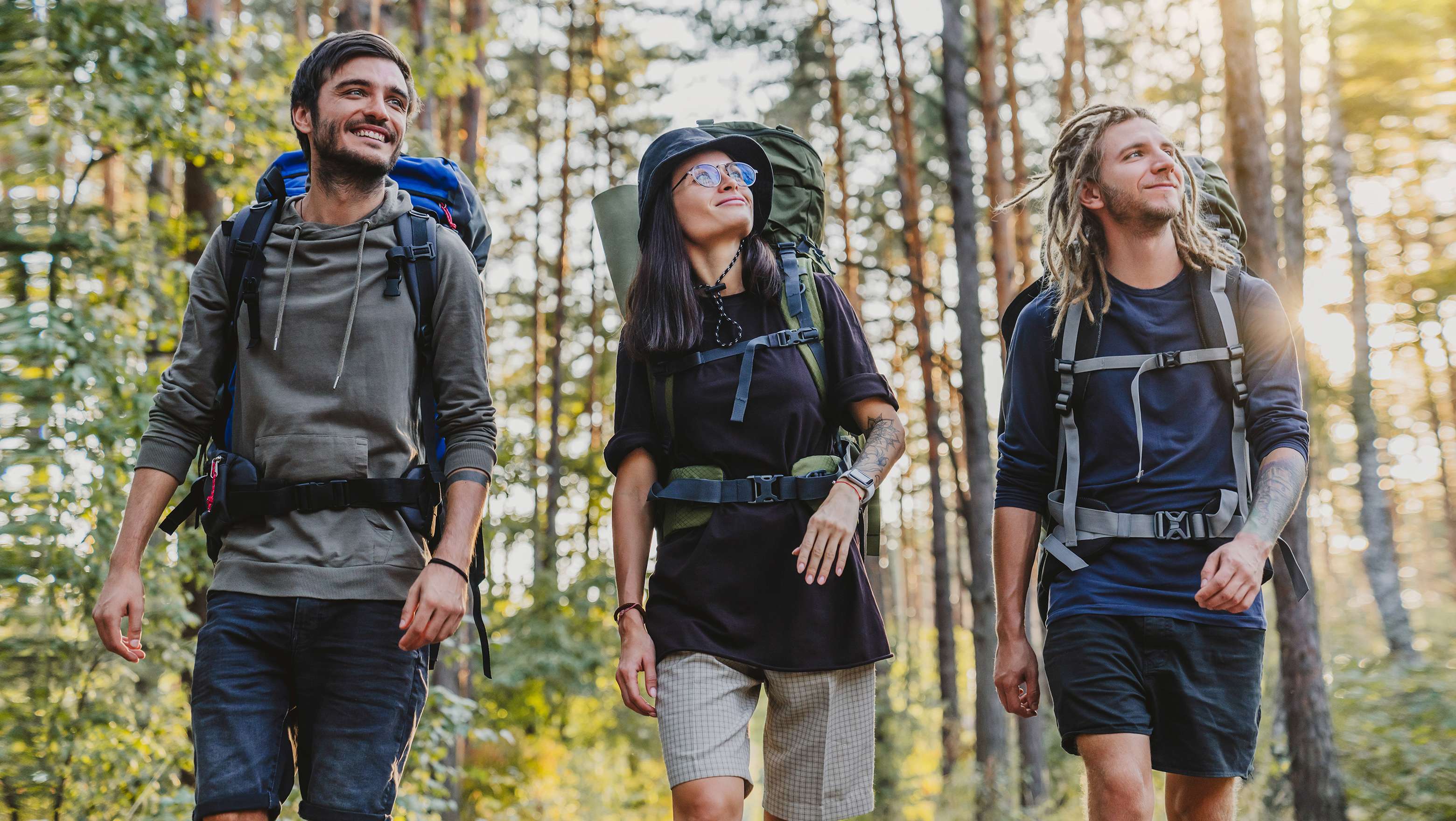 Allianz Gesundheitswelt - nachhaltige Sportausrüstung: Ein Trio aus zwei Männern und einer Frau, gekleidet mit Hut und Brille, wandern durch einen Wald. Alle drei haben einen Wanderrucksack auf dem Rücken.