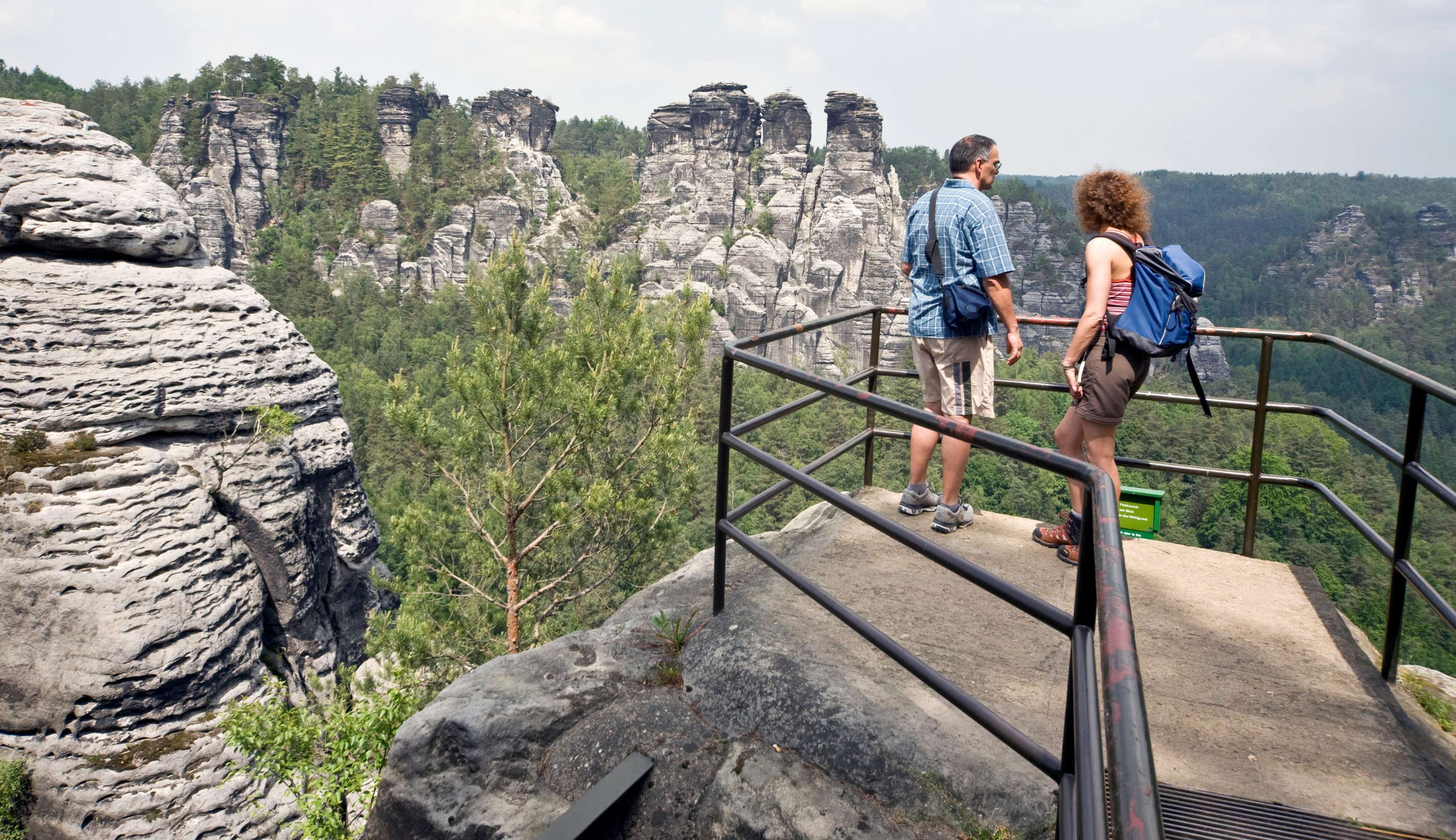 Allianz Gesundheitswelt - Urlaub vor der Haustüre: Ein älterer Mann und eine Frau stehen auf einer Aussichtsplattform. Im Hintergrund sind Gesteinsformationen und Wälder zu erkennen.