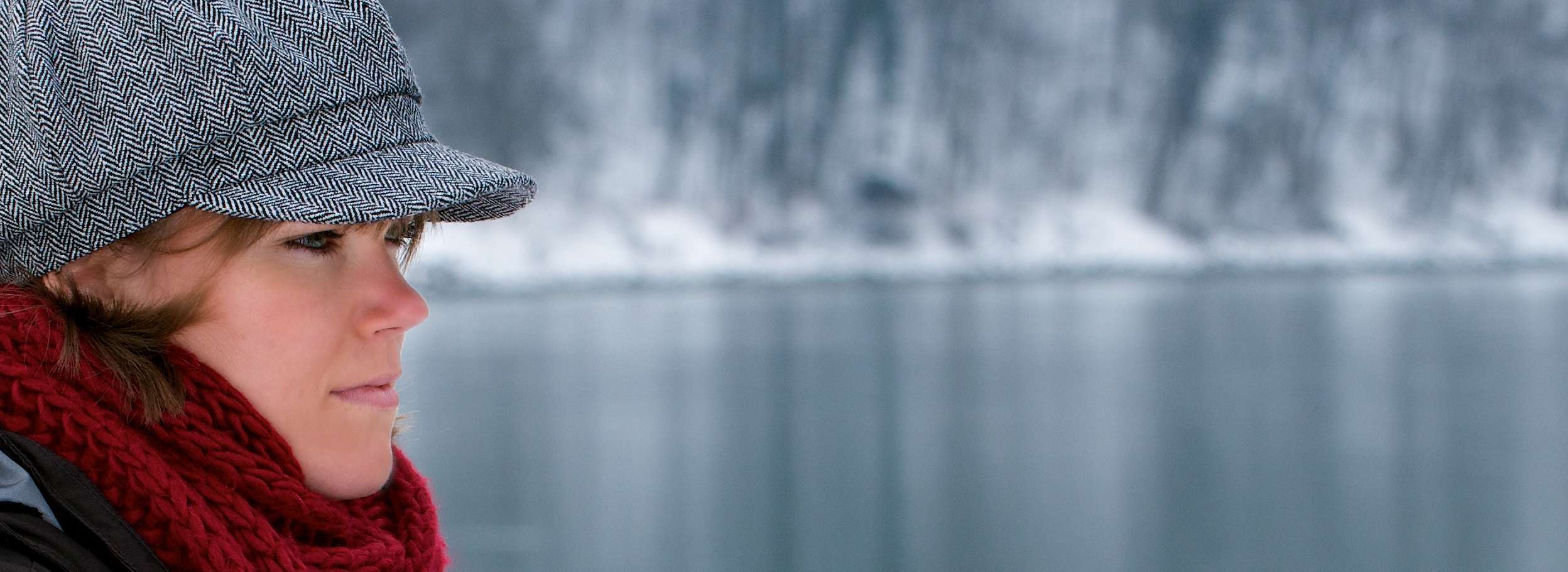 Allianz Gesundheitswelt - Feiertagsblues vorbeugen: eine junge Frau mit Winterjacke, rotem Schal und einer Kappe sitzt an einem See und blickt in die Ferne.