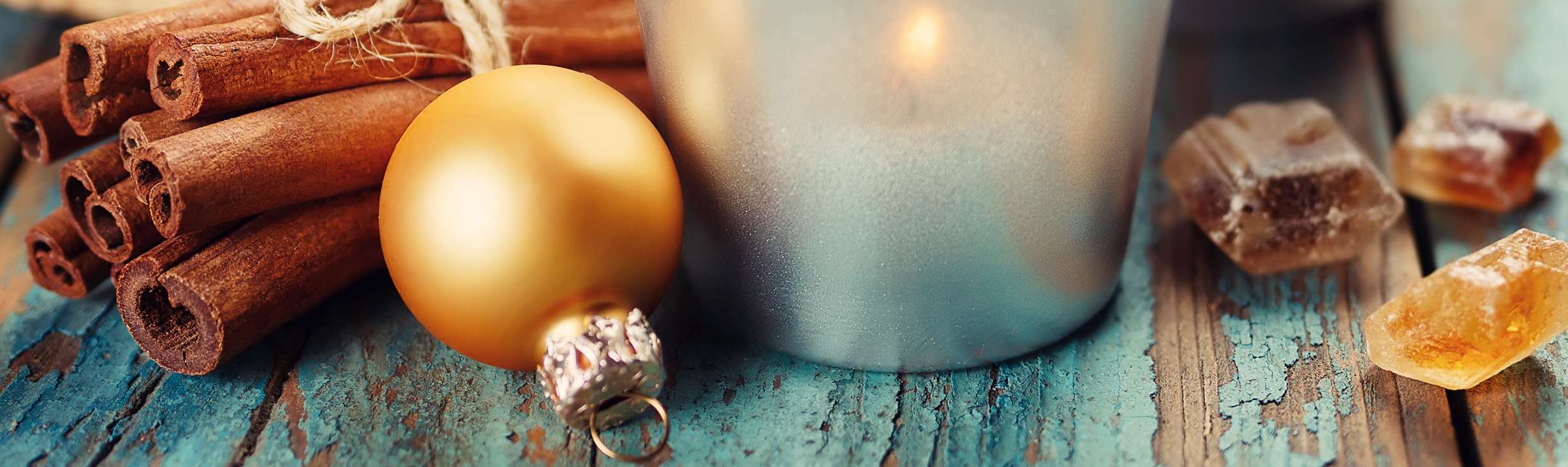 Allianz Gesundheitswelt - Stressfreie Weihnachten: ein Teelichtglas steht auf einem türkisenen Holzuntergrund. Links daneben liegen zusammengebundene Zimtstanden und eine goldene Christbaumkugel.