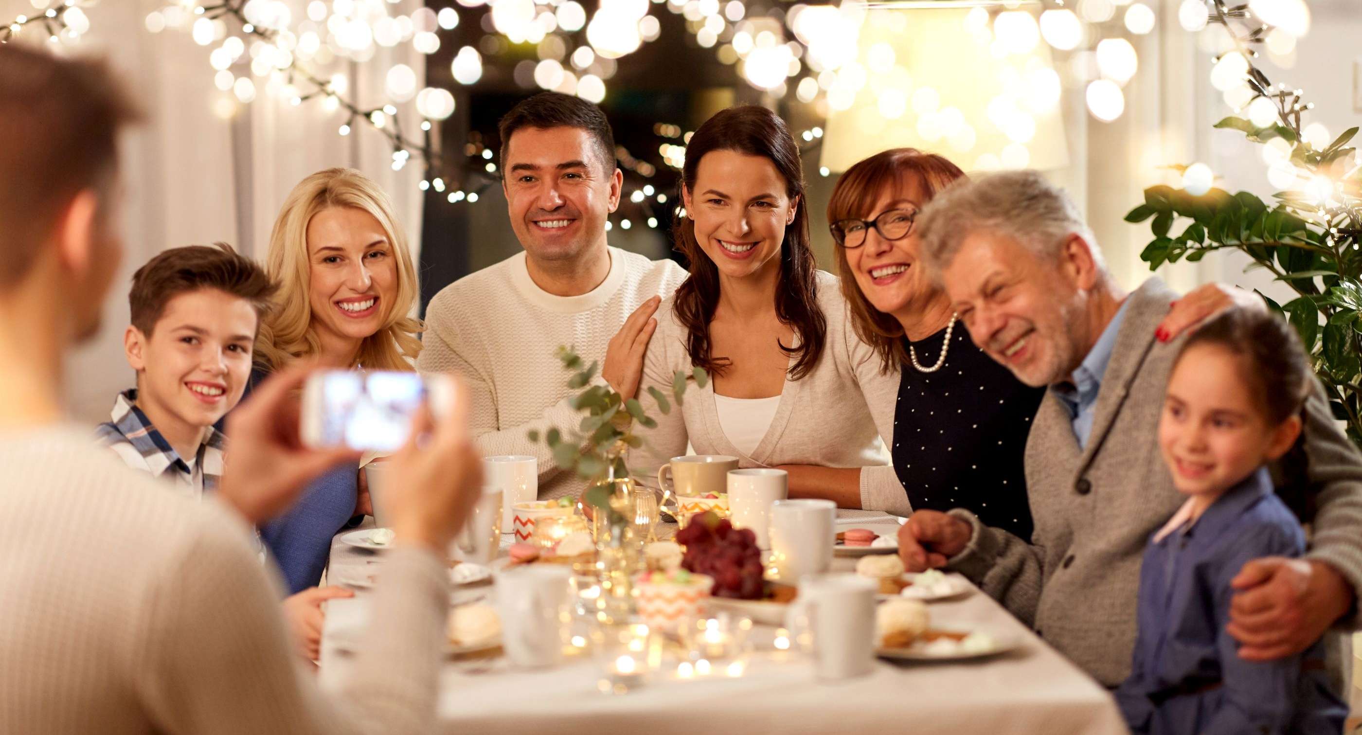 Allianz Gesundheitswelt - Stressfreie Weihnachten: eine Familie sitzt lächelnd an einem gedeckten Tisch, während eine Person ein Foto von ihnen macht. Im Hintergrund leuchten Lichterketten.