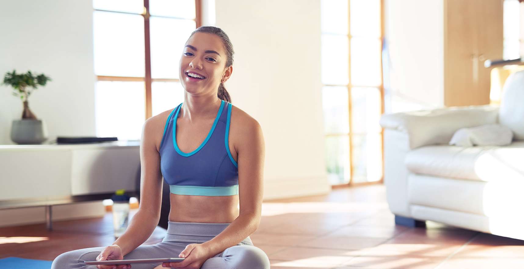 Allianz Gesundheitswelt - Physiotherapie online: eine junge Frau mit zusammengebundenen Haaren sitzt im Schneidersitz auf einer Fitnessmatte.