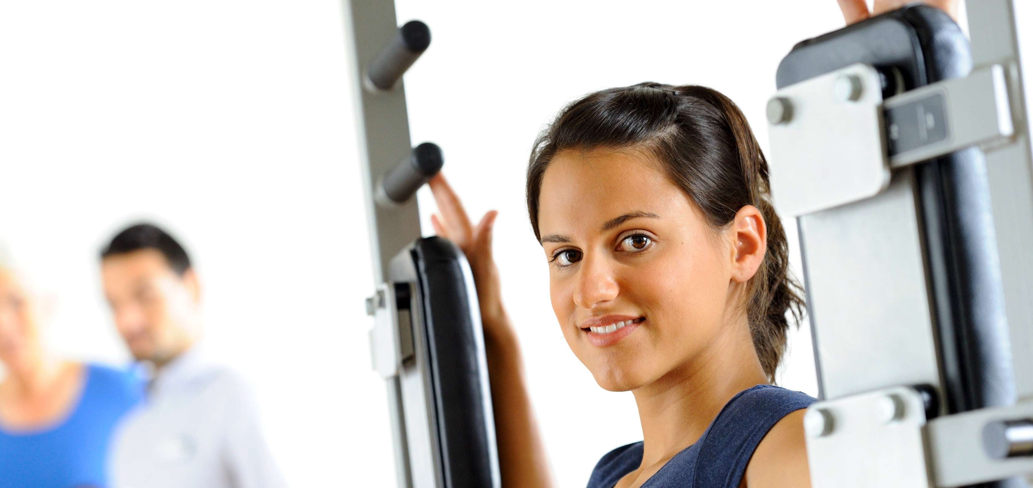 Allianz Gesundheitswelt - Professionelles Rückentraining: eine junge Frau mit blauem Sportoberteil trainiert an einem Fitnessgerät ihren Rücken.