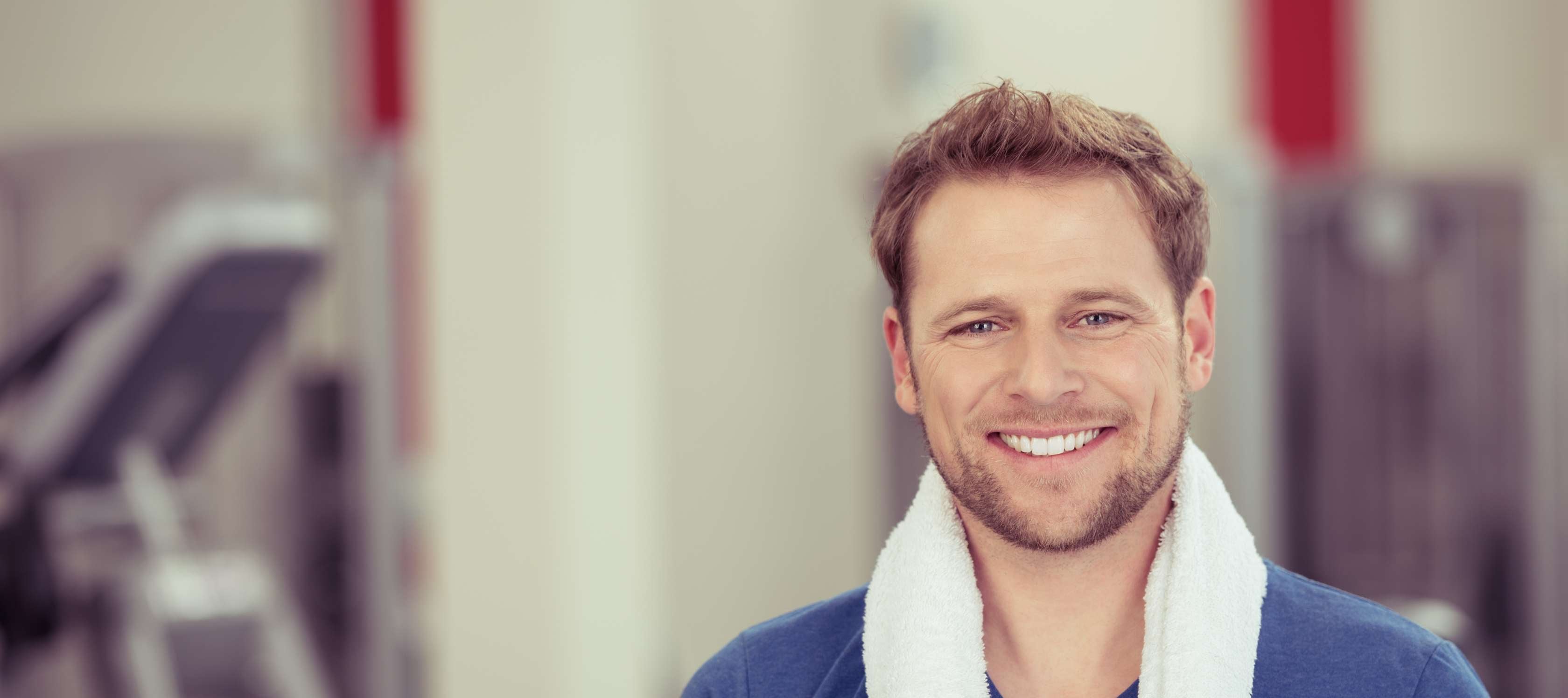 Allianz Gesundheitswelt - Reha-Vermittlung: ein Mann mit blauem T-Shirt und einem weißen Handtuch um die Schultern  ist im Gym und lacht in die Kamera.