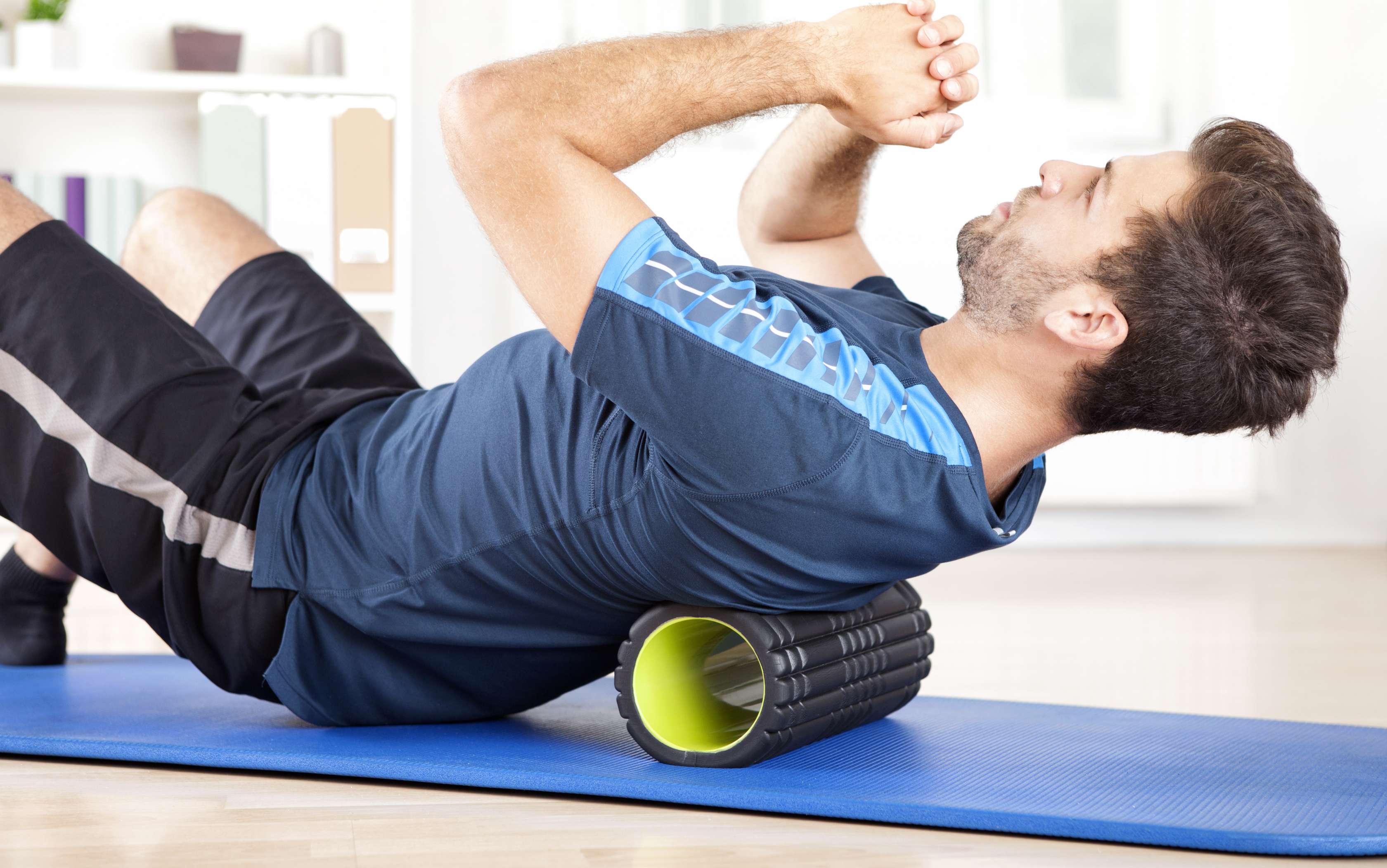 Allianz Gesundheitswelt - Geräte für das Training Daheim: Ein Mann mit dunkelblauem Oberteil liegt mit seinem Rücken auf einer Faszienrolle und hält sich mit ausgewählten Übungen zuhause fit.