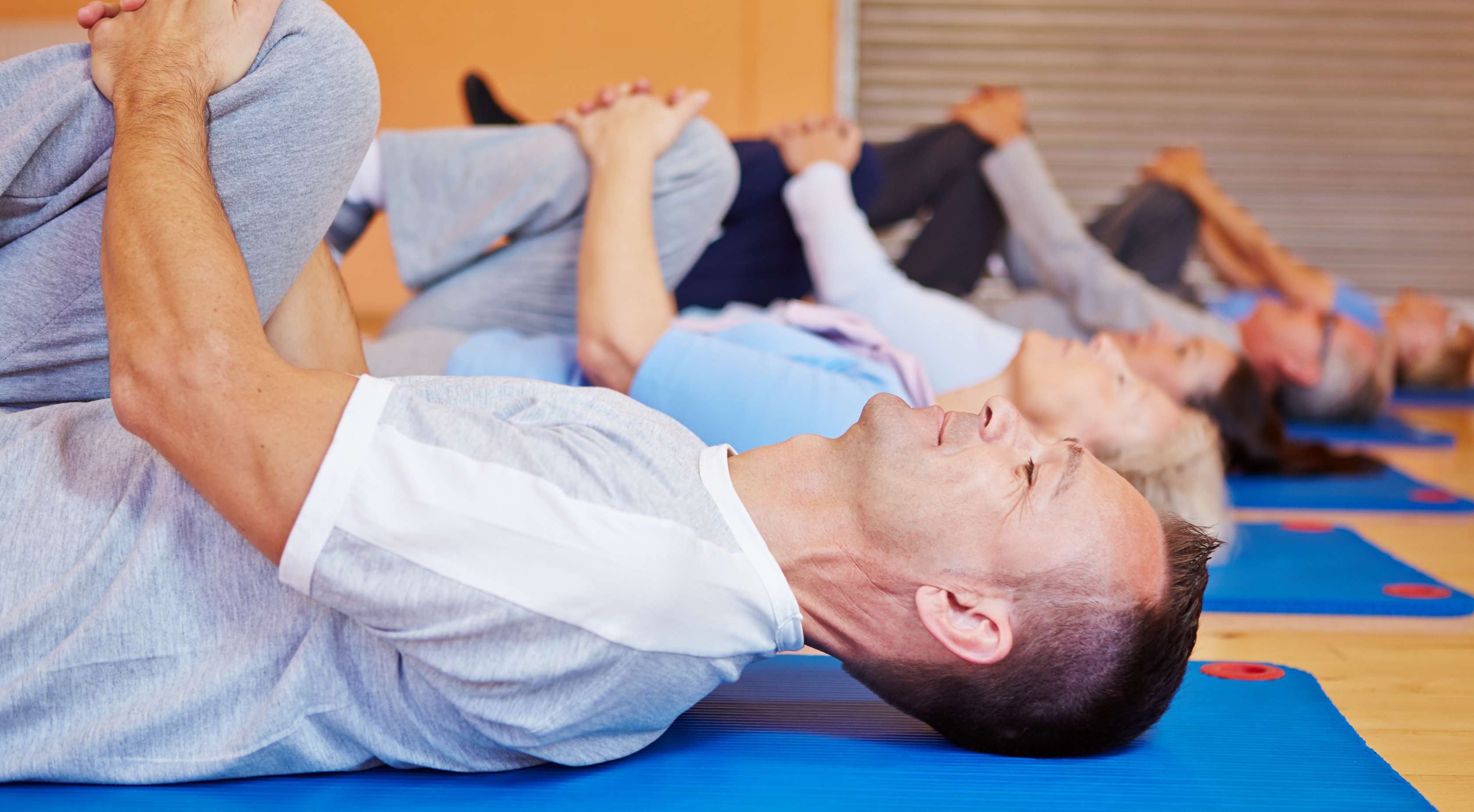 Allianz Gesundheitswelt - Rückentraining: Eine Gruppe von Menschen liegt in einer Turnhalle auf Fitnessmatten und zieht ein Knie in Richtung Kinn, um den Rücken zu entlasten.